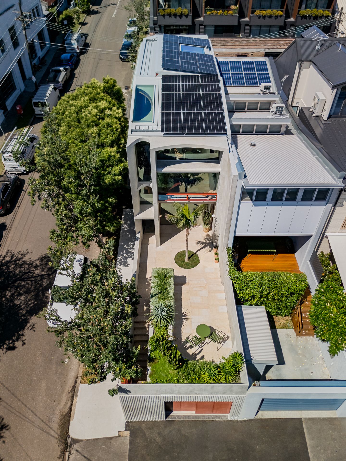 Aerial view of a modern concrete townhouse with rooftop solar panels, a landscaped courtyard, and a palm tree on a tree-lined street.