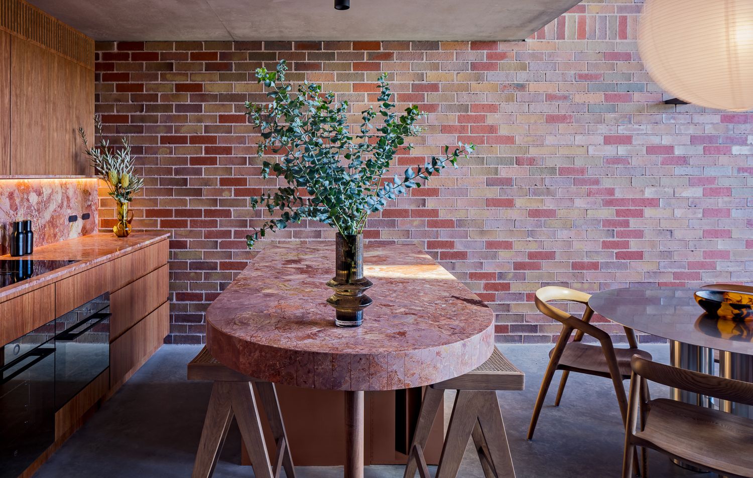 Pink stone dining table on trestle legs with eucalyptus in a vase, set against a warm multicolored brick wall.