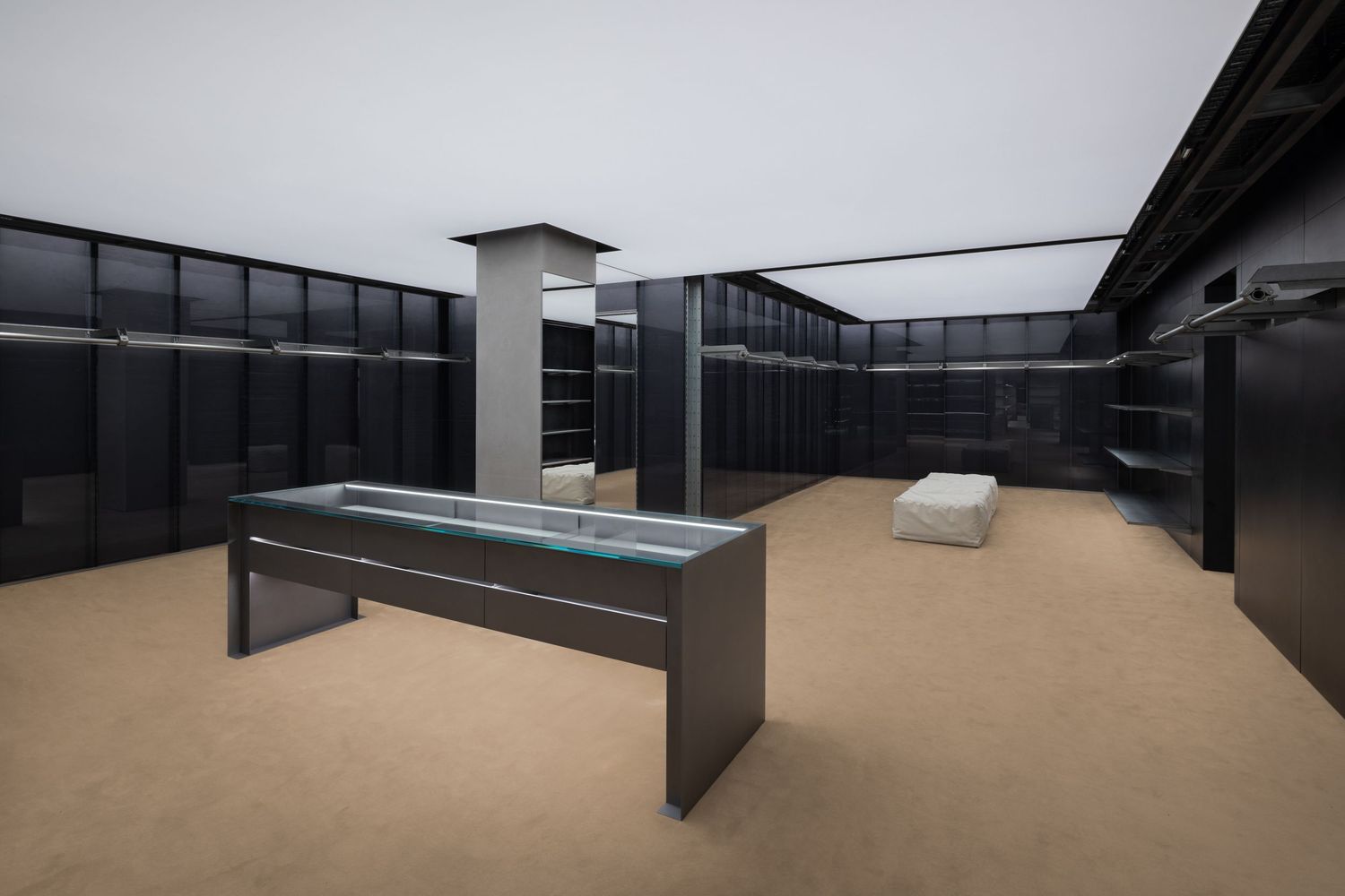 Spacious luxury boutique interior with black wall shelving, a glass-topped display table, a white bench, and beige carpet flooring.