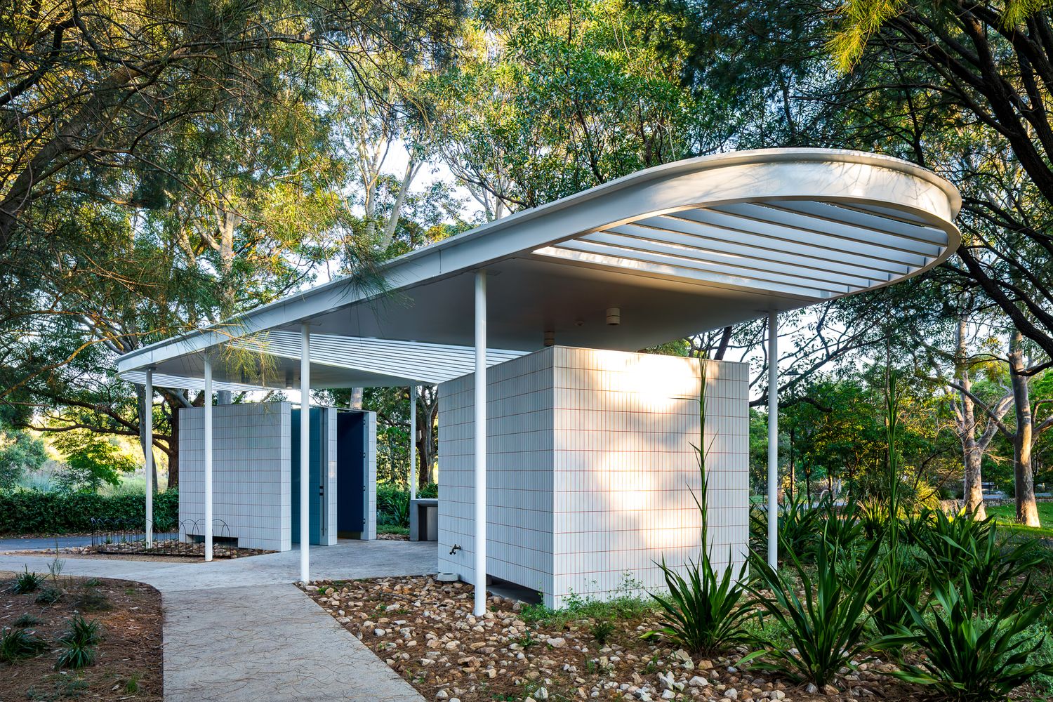 Modern white park amenity building with a curved slatted roof canopy, blue doors and native grasses set among shady trees.