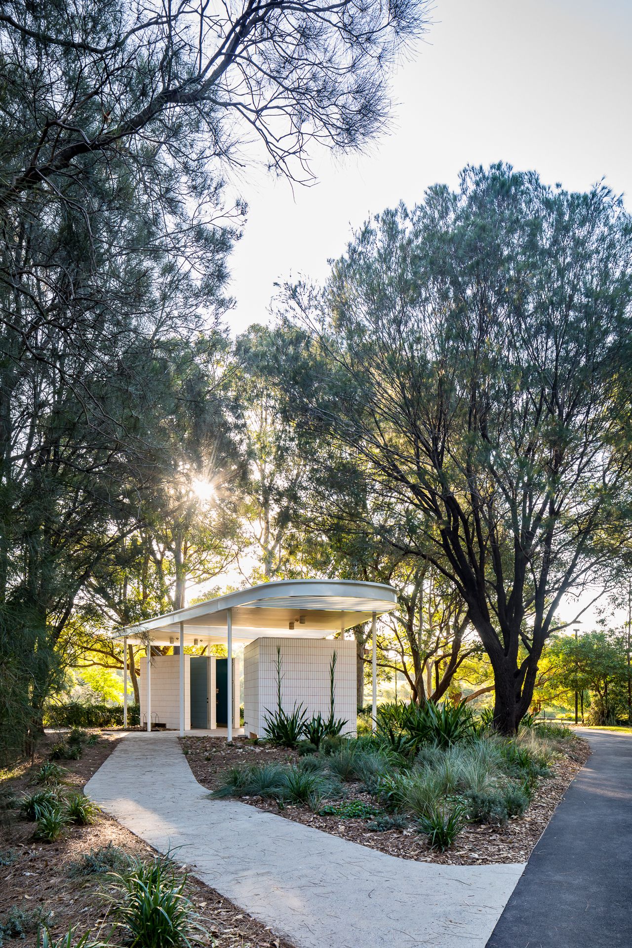 A curved concrete path leads through bushland to a small white park building with a sleek canopy roof as the sun sets behind trees.