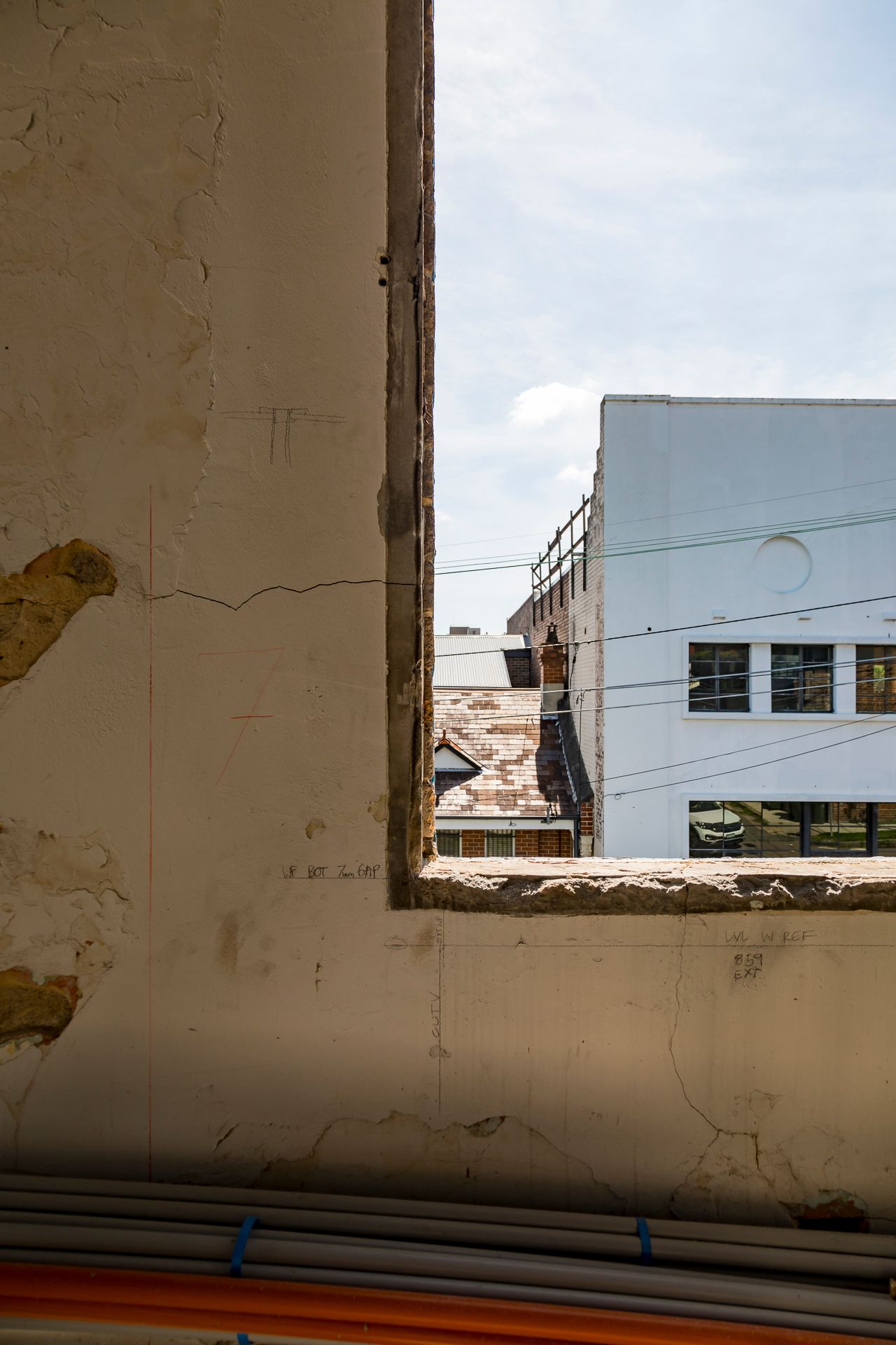 View through a rough window opening in a weathered plaster wall toward a white building and a damaged rooftop under a blue sky.
