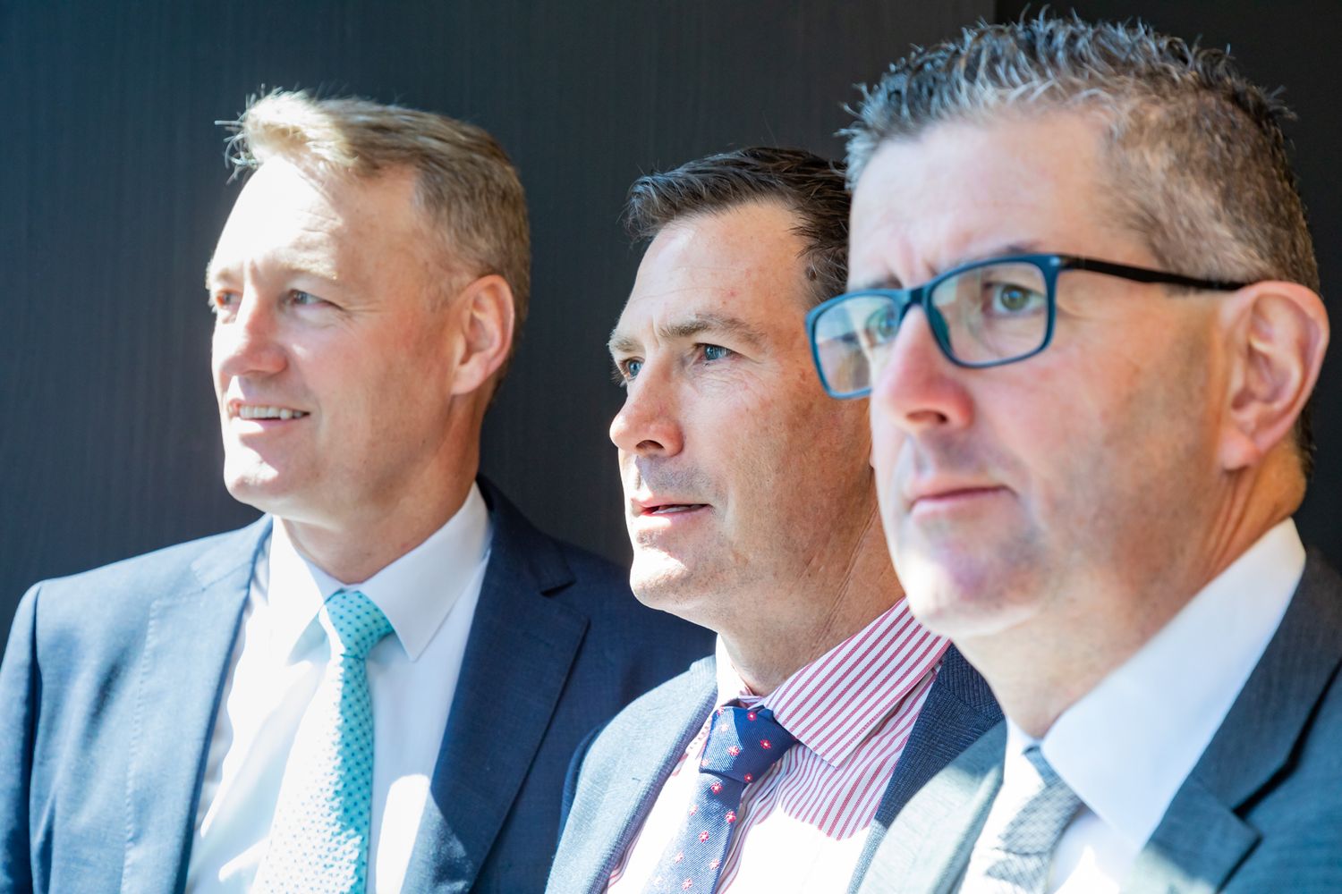 Three men in business suits, one wearing glasses, look off to the side in a close-up professional group portrait against a dark wall.