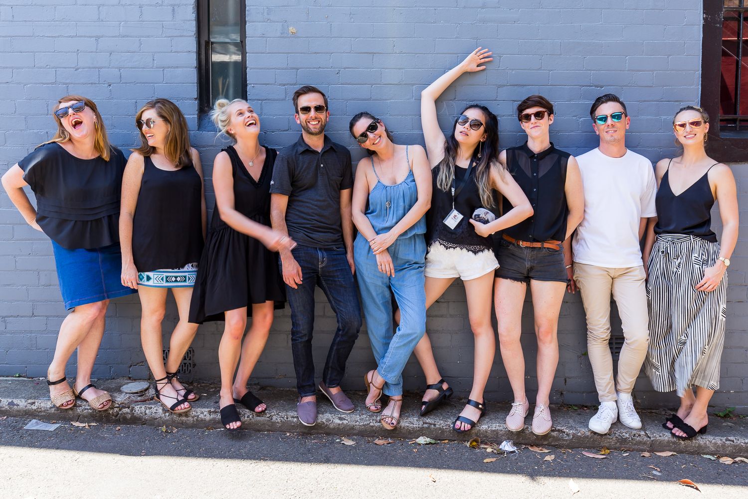 Group of nine friends and colleagues posing playfully in a row against a gray brick wall on a sunny day, laughing and striking fun poses.