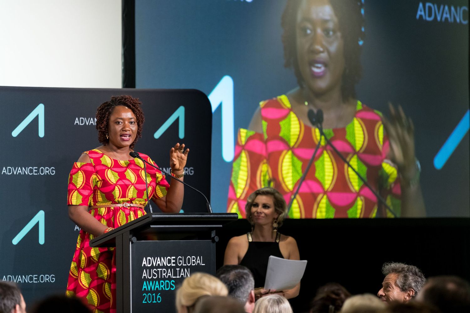 Woman in a bright patterned dress speaks at the Advance Global Australian Awards 2016 podium while a seated woman looks on.