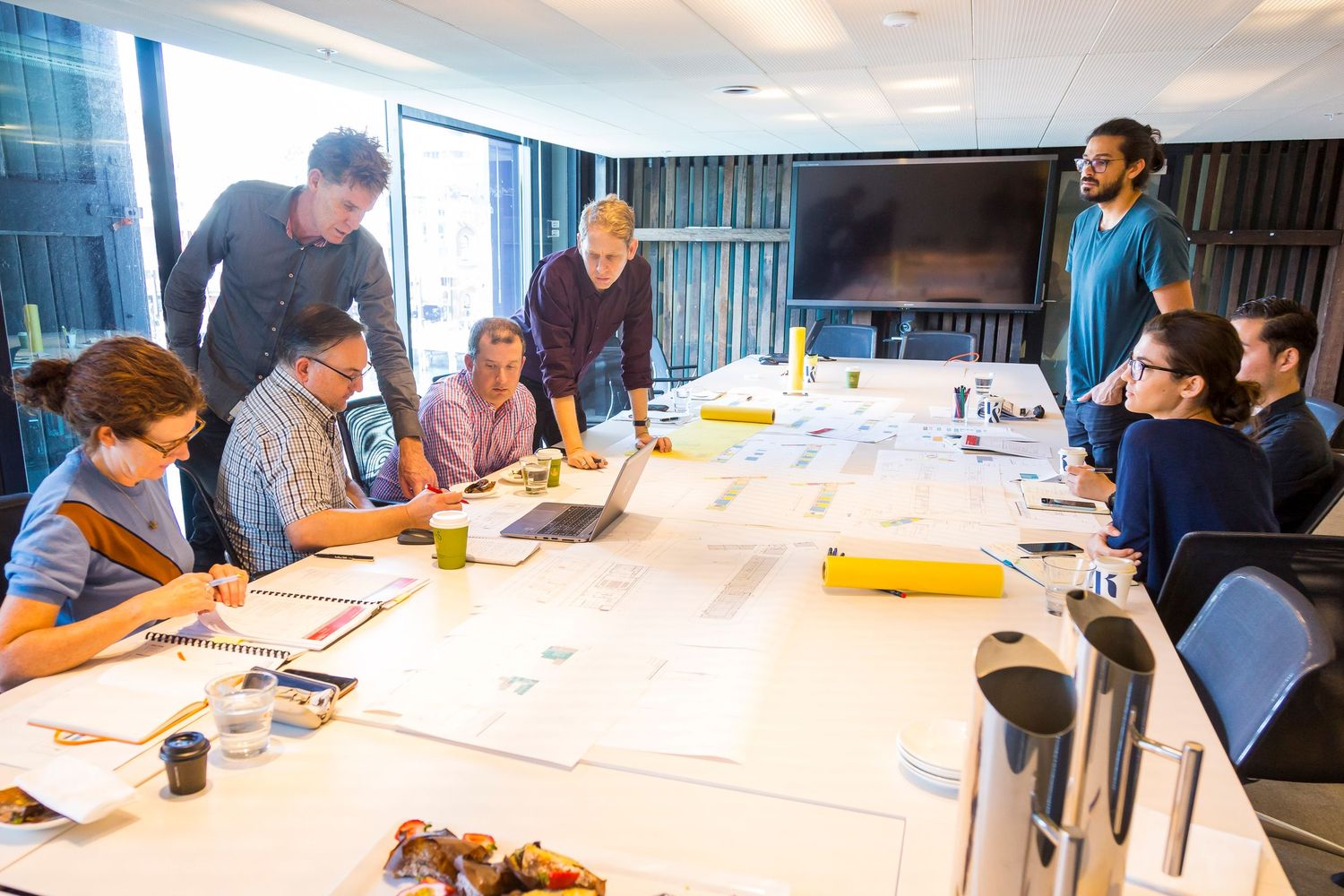 A team of people gathers around a large meeting table covered with plans and documents, collaborating in a bright modern office.