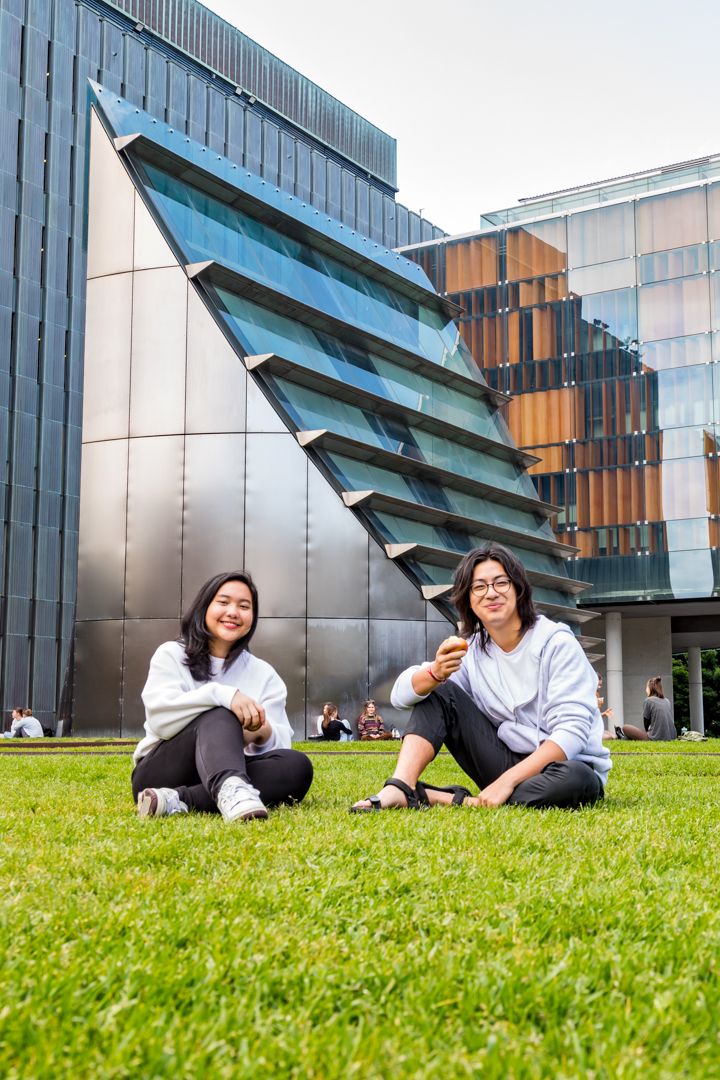 Two smiling young women in white tops sit on a grassy lawn in front of modern glass and steel university buildings.
