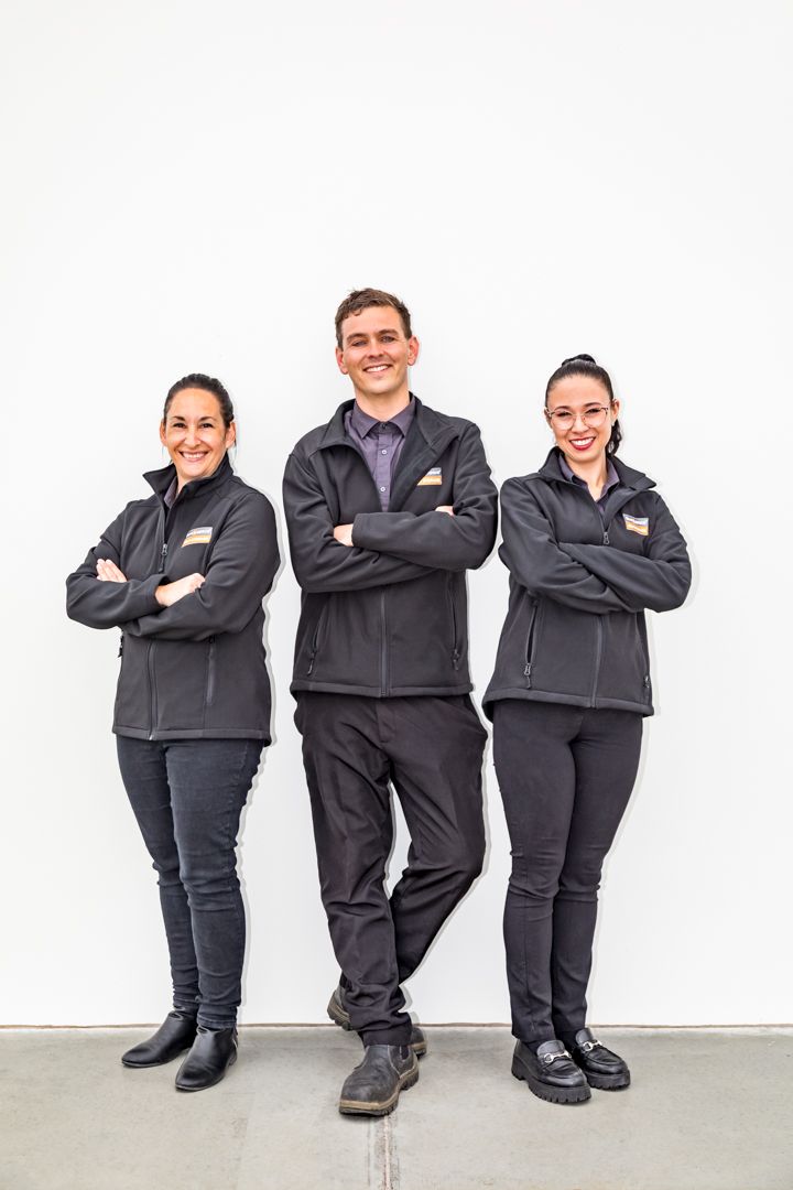 Three smiling staff members in matching dark uniform jackets stand with arms crossed against a plain white wall.