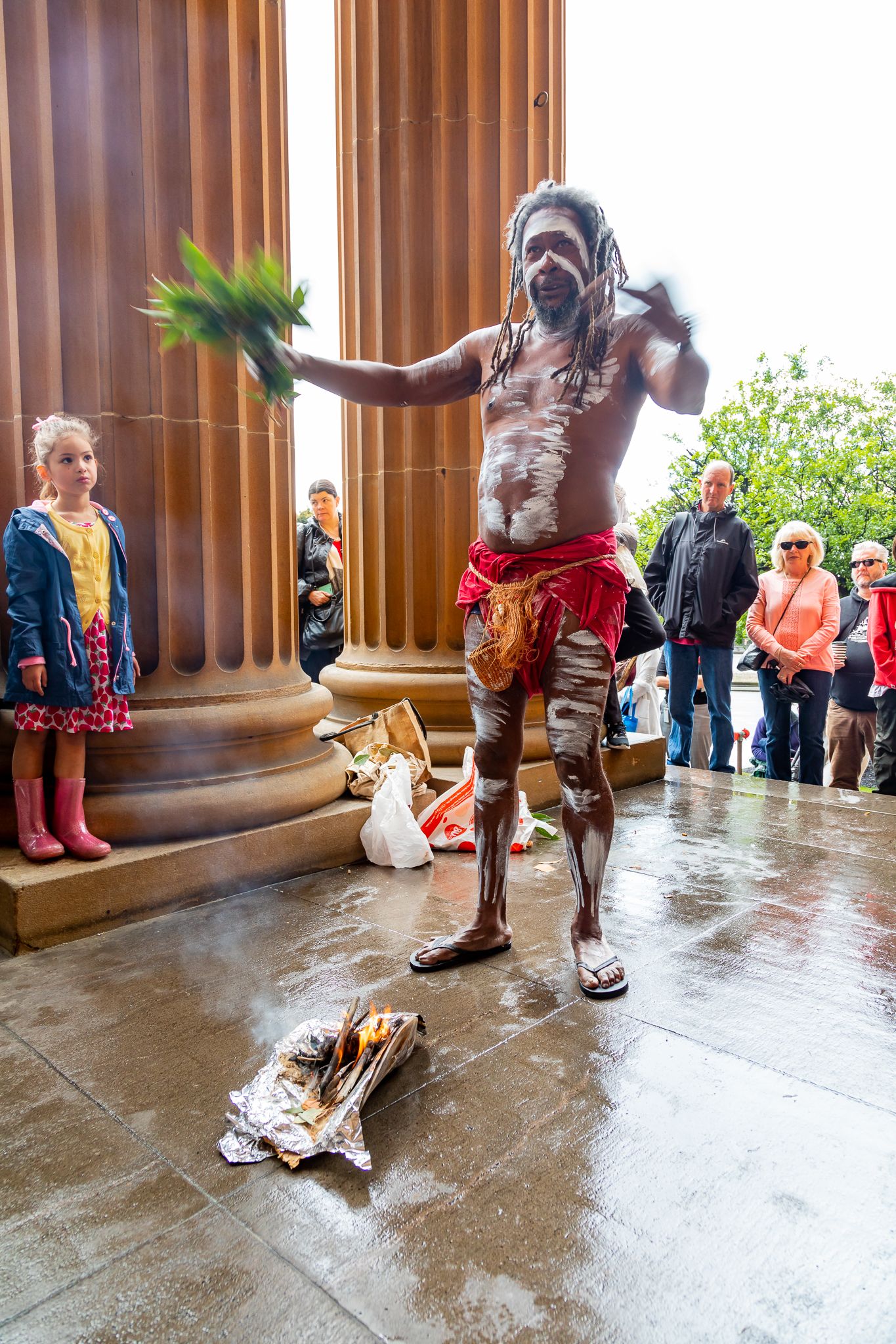 Man with body paint and red cloth holds green leaves over a smoking ceremony fire beside stone columns as a girl and onlookers watch.