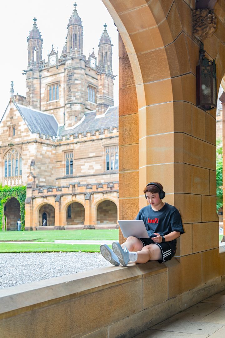 Student wearing headphones sits on a sandstone ledge working on a laptop beside a historic Gothic university building.