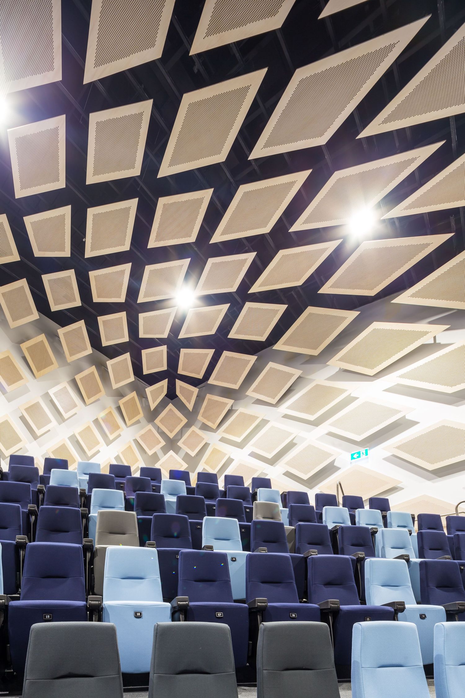 Auditorium ceiling with diamond-shaped acoustic panels in a geometric lattice above rows of blue and grey theatre seats.