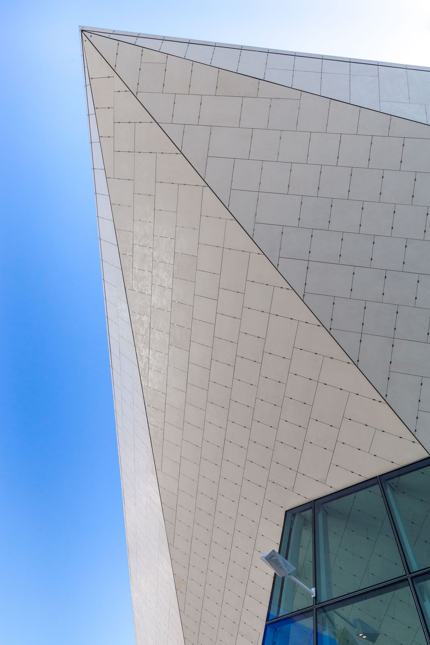 Close-up of a sharply angled white tiled building corner rising against a blue sky beside a glass window section.