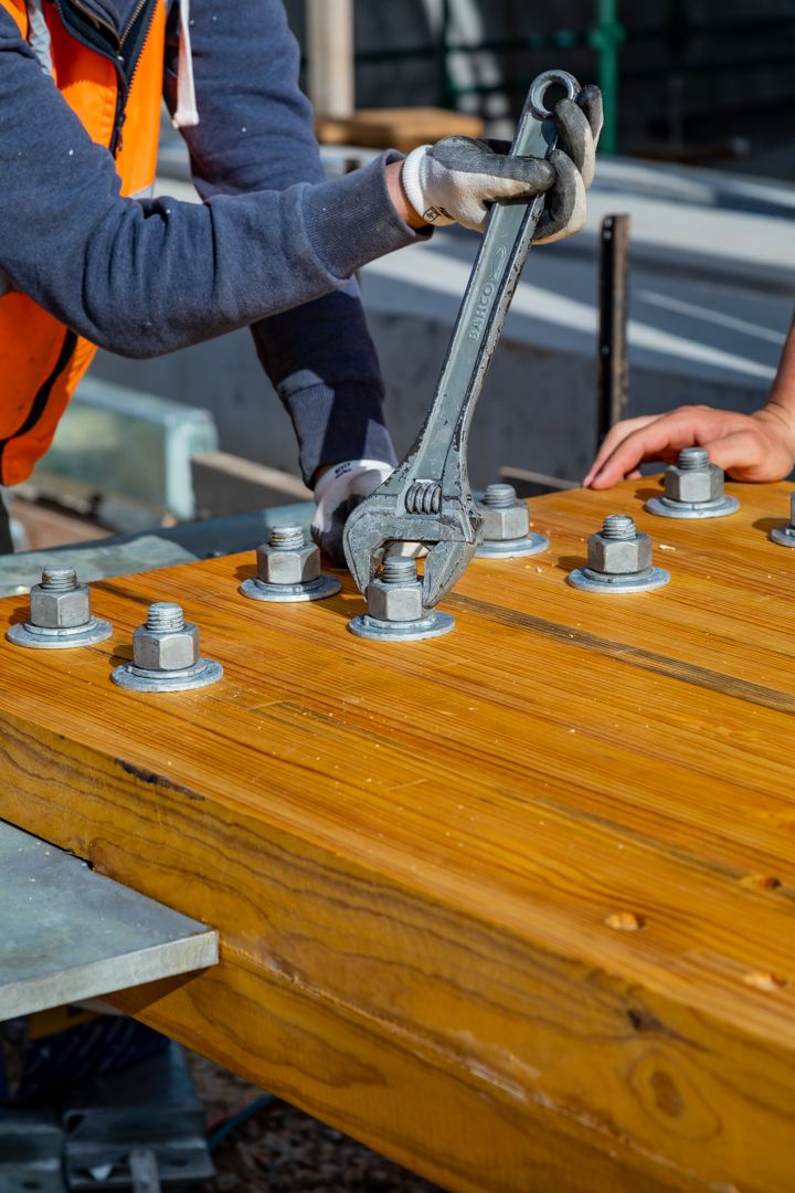 Worker tightening large steel bolts and nuts on a heavy timber beam with a wrench at a construction site.