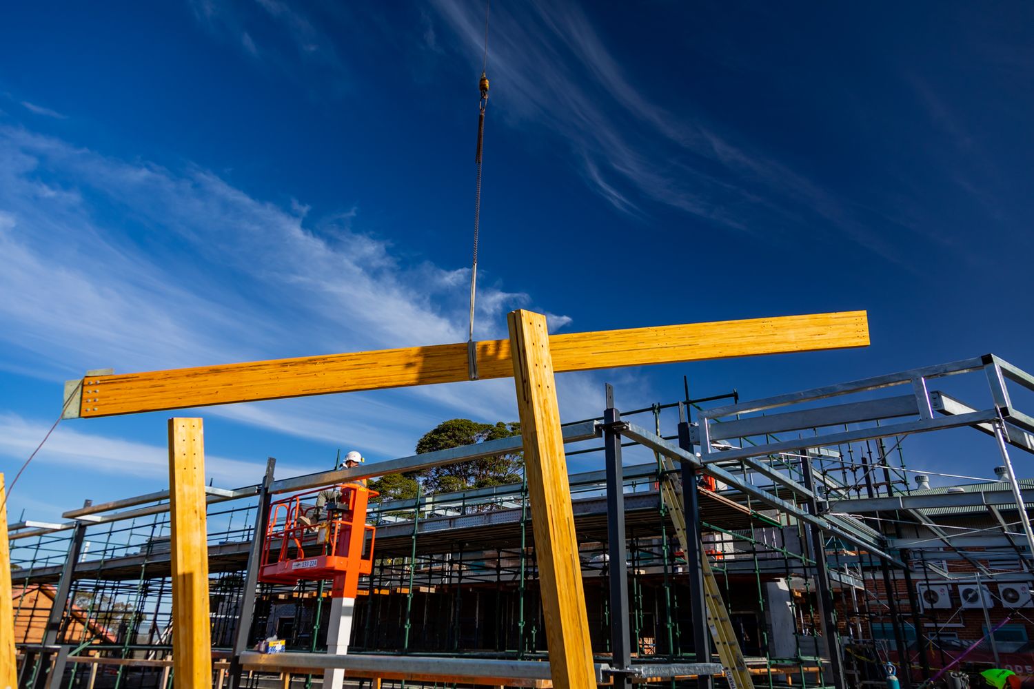 Large timber beams frame a blue sky above a multistorey construction site with scaffolding and a crane cable.