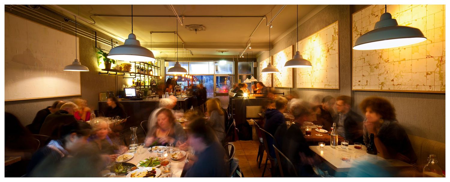 Cozy busy restaurant with diners at tables under pendant lamps, warm lighting and map-style artwork on the walls.