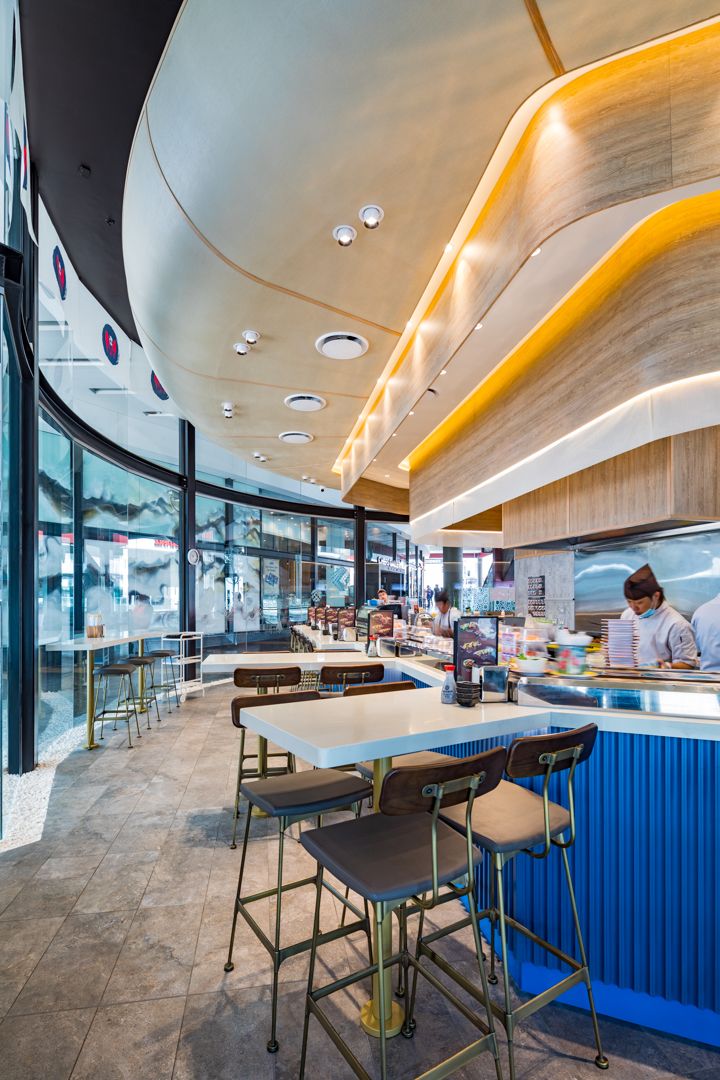 Curved restaurant interior with warm wood ceiling, gold lighting, blue-fronted counter, bar stools and chefs preparing food.