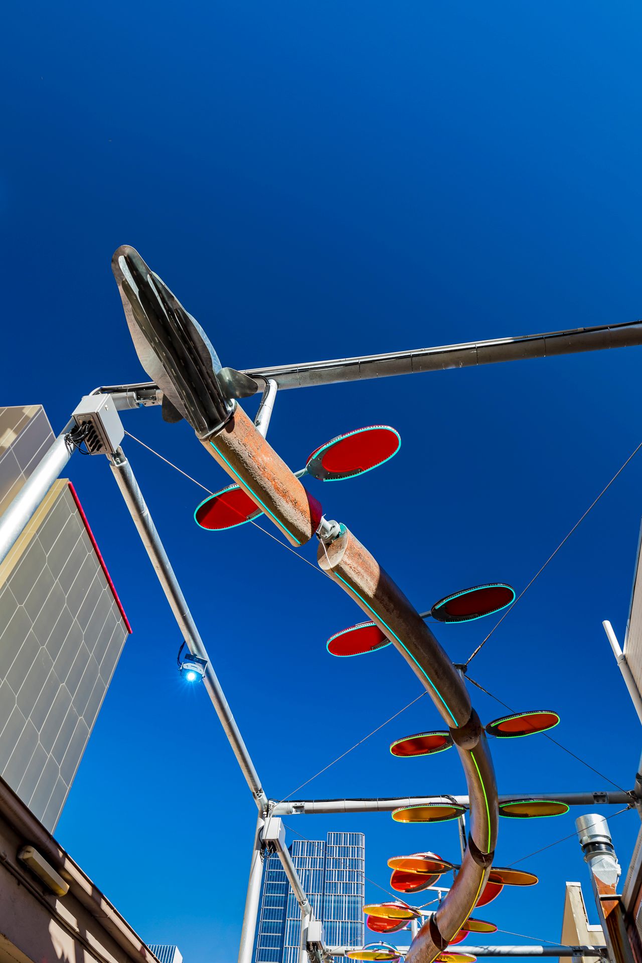 Low-angle view of a suspended kinetic sculpture with red oval discs against a deep blue sky and city buildings.