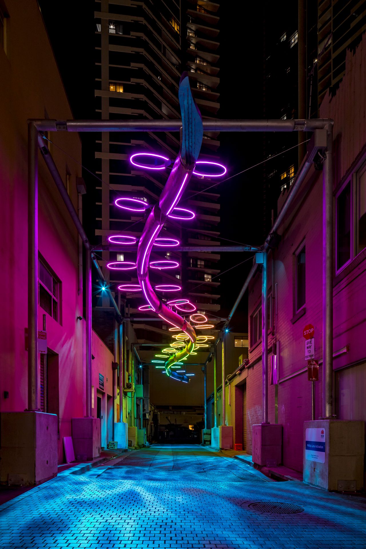 A purple neon spiral sculpture with yellow-green accents hangs above a pink-toned alley at night in the city.