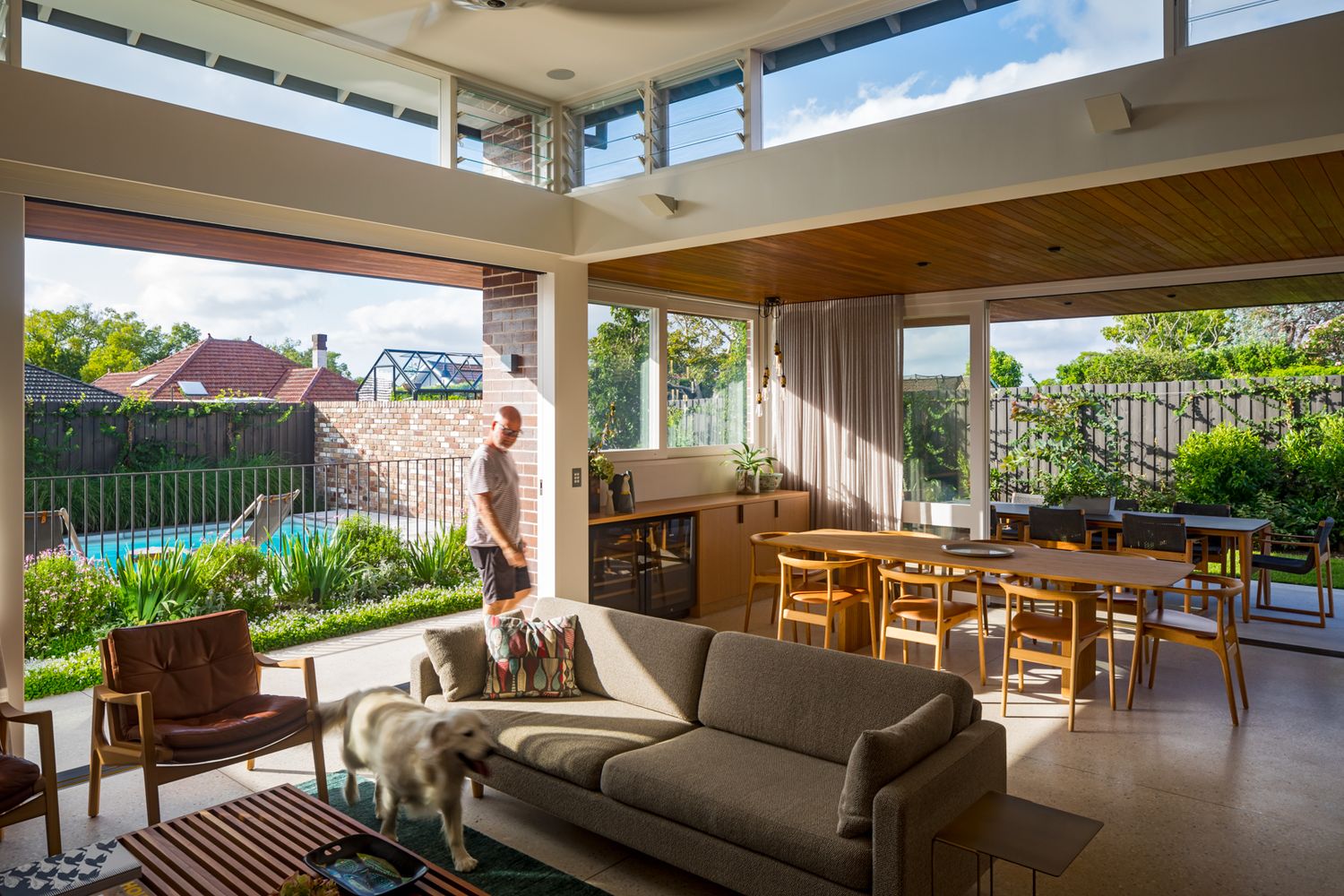 Open living and dining area with a timber ceiling and sliding doors leading to a leafy garden, with a dog on the sofa.