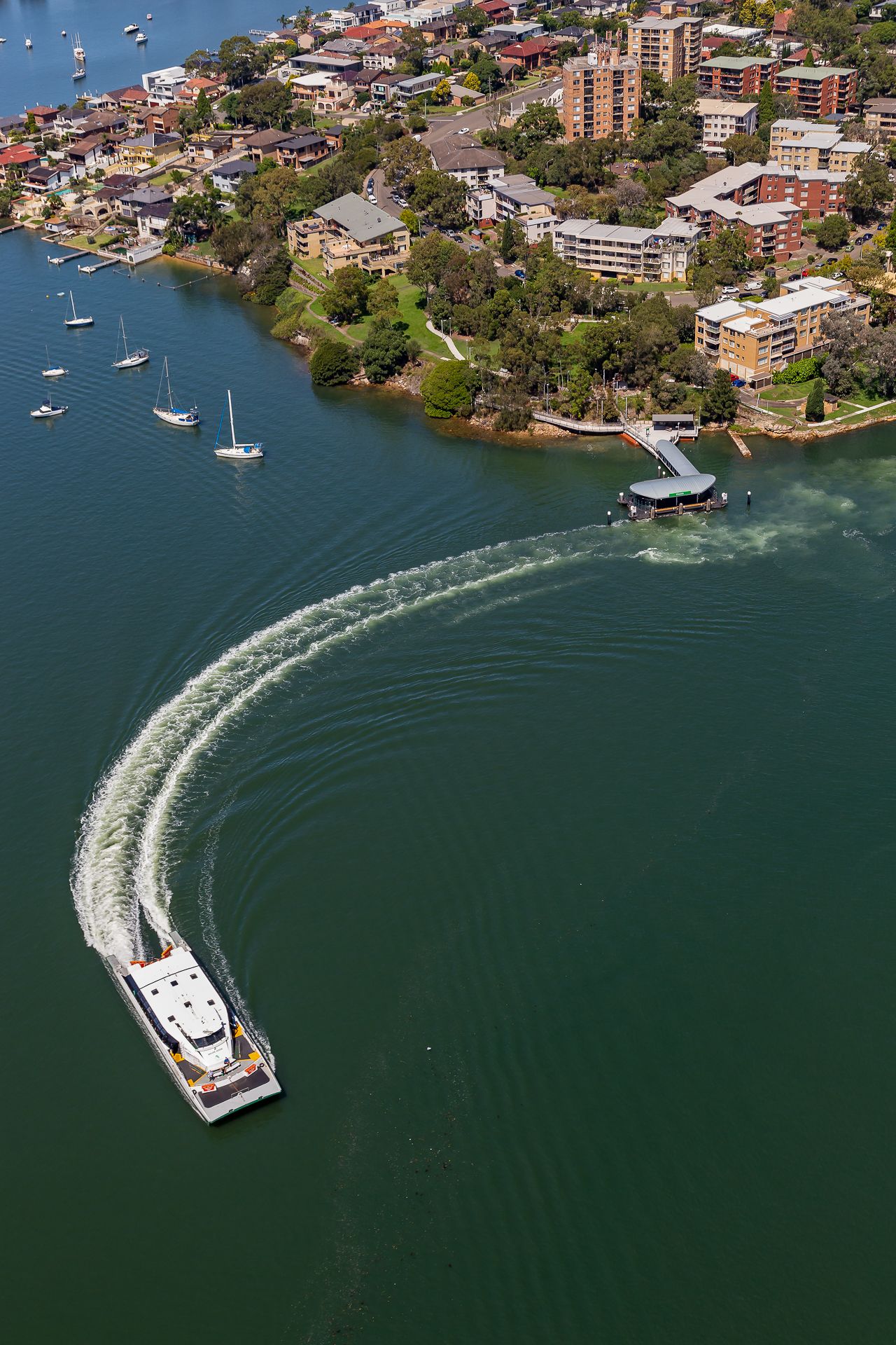 Aerial view of a ferry carving a curved wake across blue harbour water beside waterfront apartments, green parkland and moored sailboats.