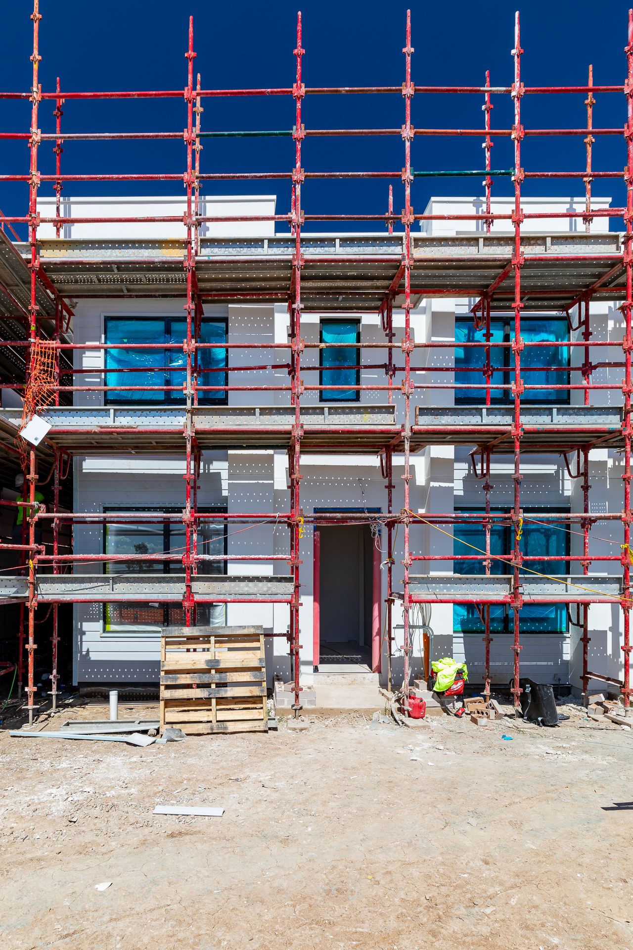 Two-storey townhouse under construction wrapped in red scaffolding with new windows, a doorway, and a worker below.