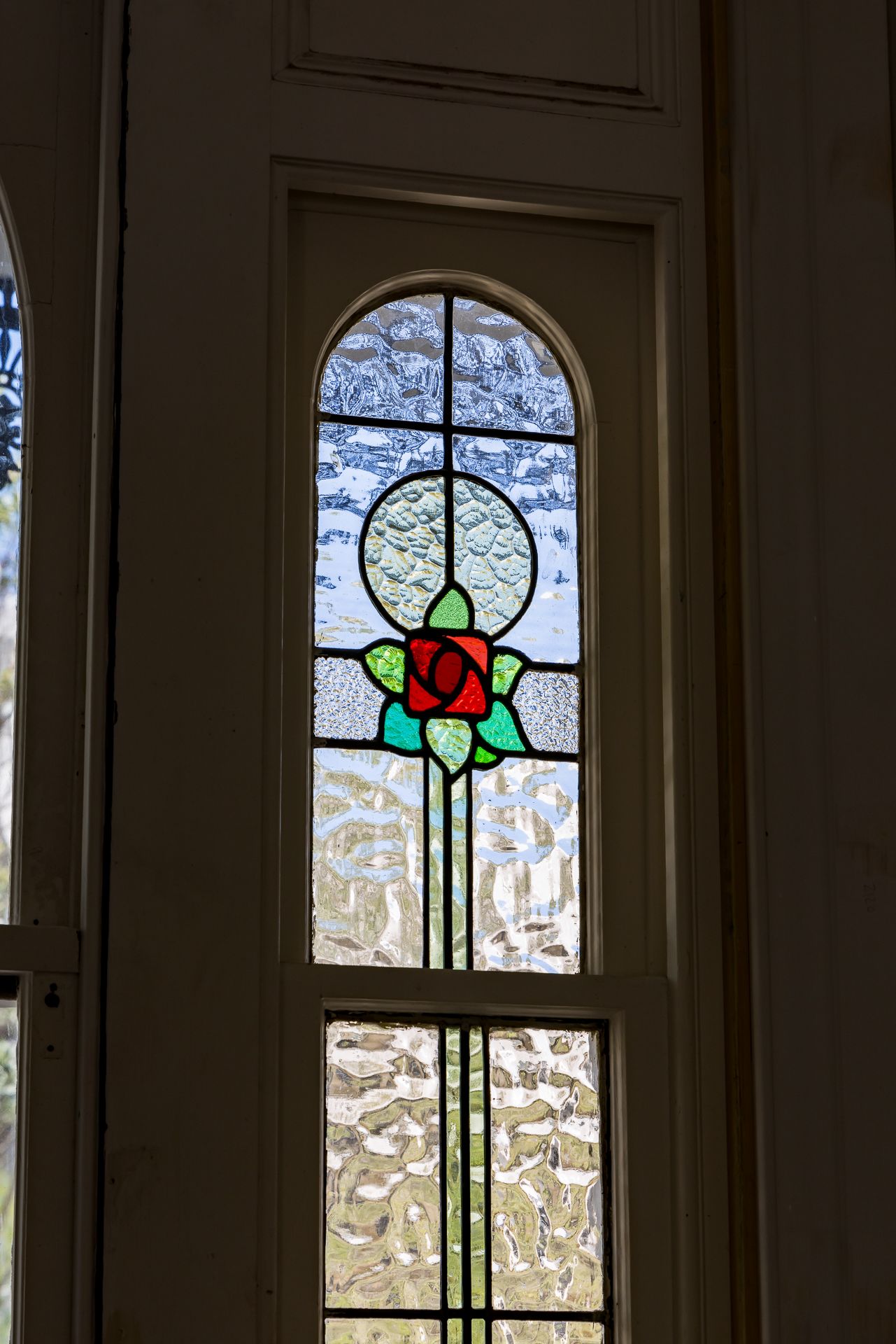 Arched stained glass window with a red rose and green leaves set in textured glass panes within a white timber frame.