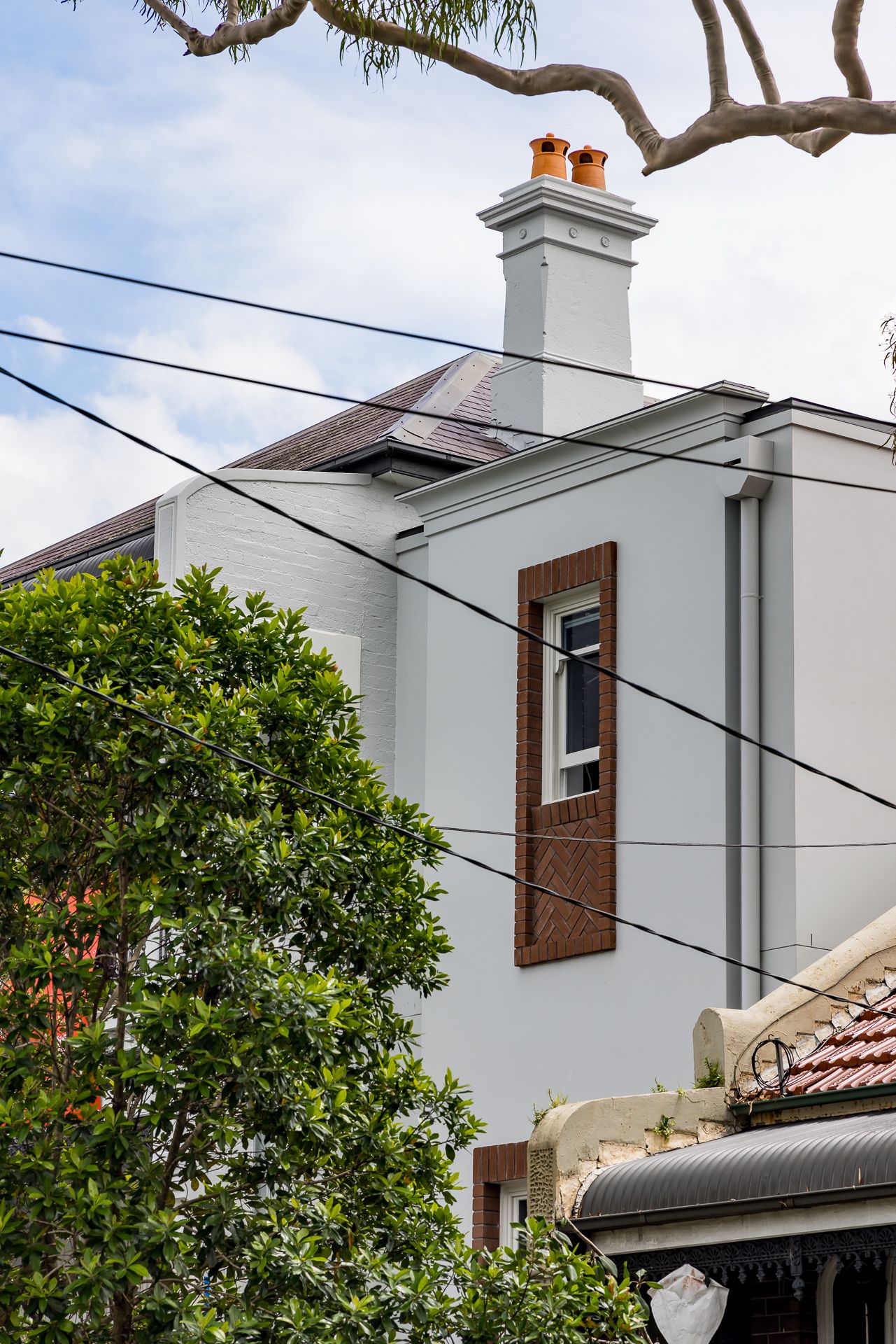 Two-storey rendered home with a brick-framed window, chimney with terracotta pots, tiled roof and green foliage in front.