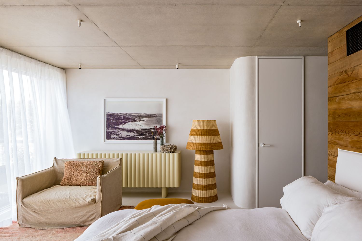 Minimalist bedroom with concrete ceiling, striped floor lamp, cream armchair, radiator cabinet and white linen bedding.