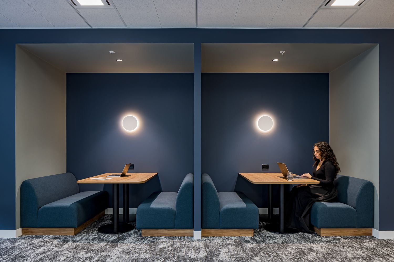 Woman working on a laptop in a navy blue office booth with banquette seating, wooden table and round wall lighting.
