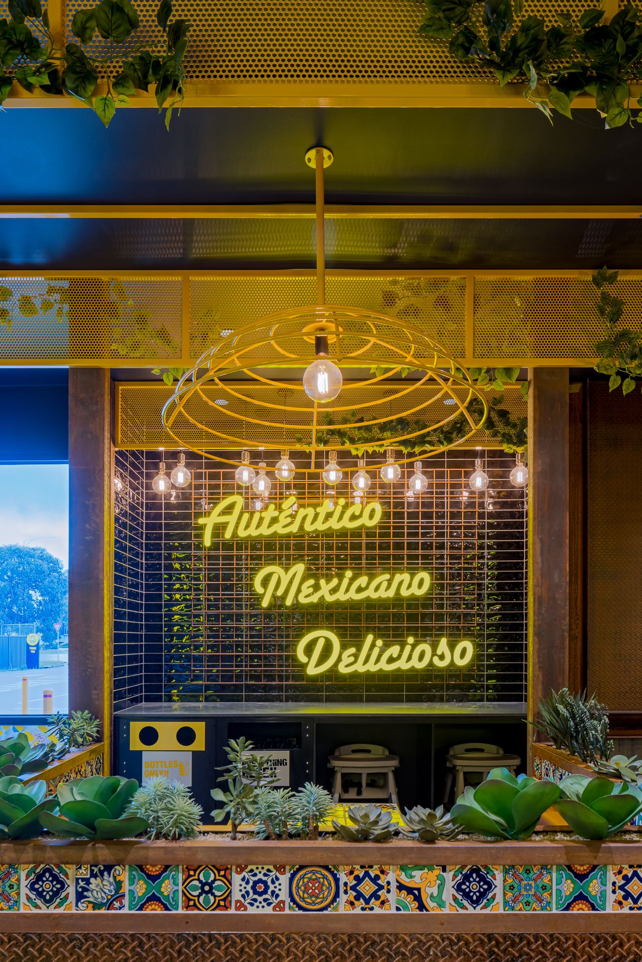 Glowing neon sign reading Auténtico Mexicano Delicioso above succulent planters with colourful Mexican tile detail.