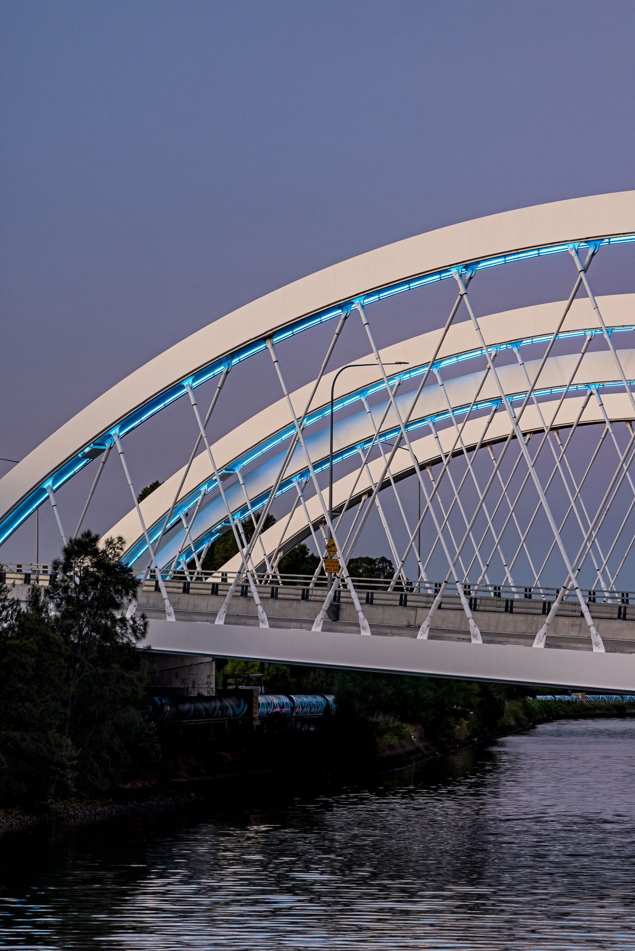 Close view of white steel bridge arches with blue LED lighting and slender cable supports above a quiet river.