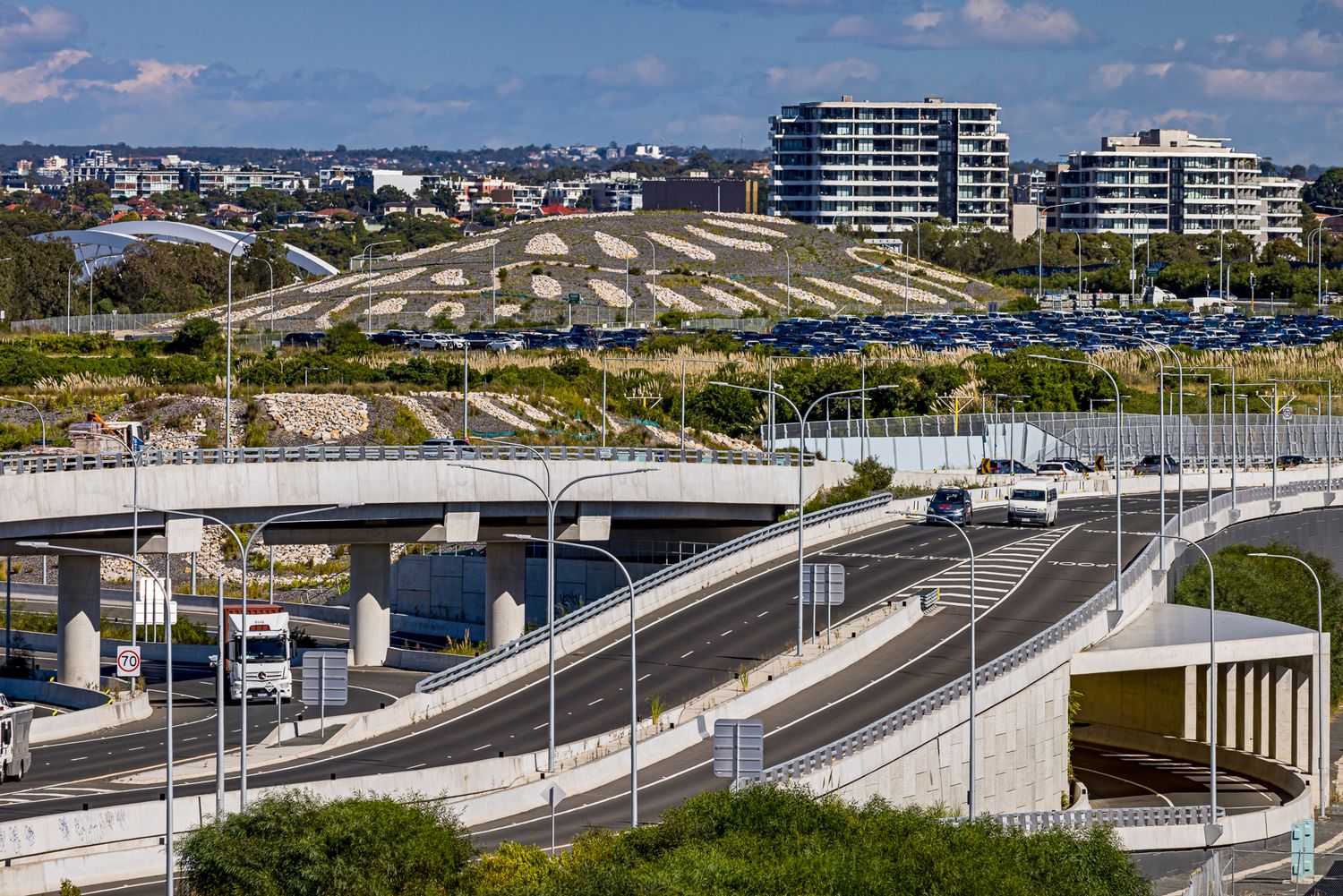 Multi-lane highway curves beneath a bridge with modern apartment buildings, green hills, and parked cars under a partly cloudy sky.