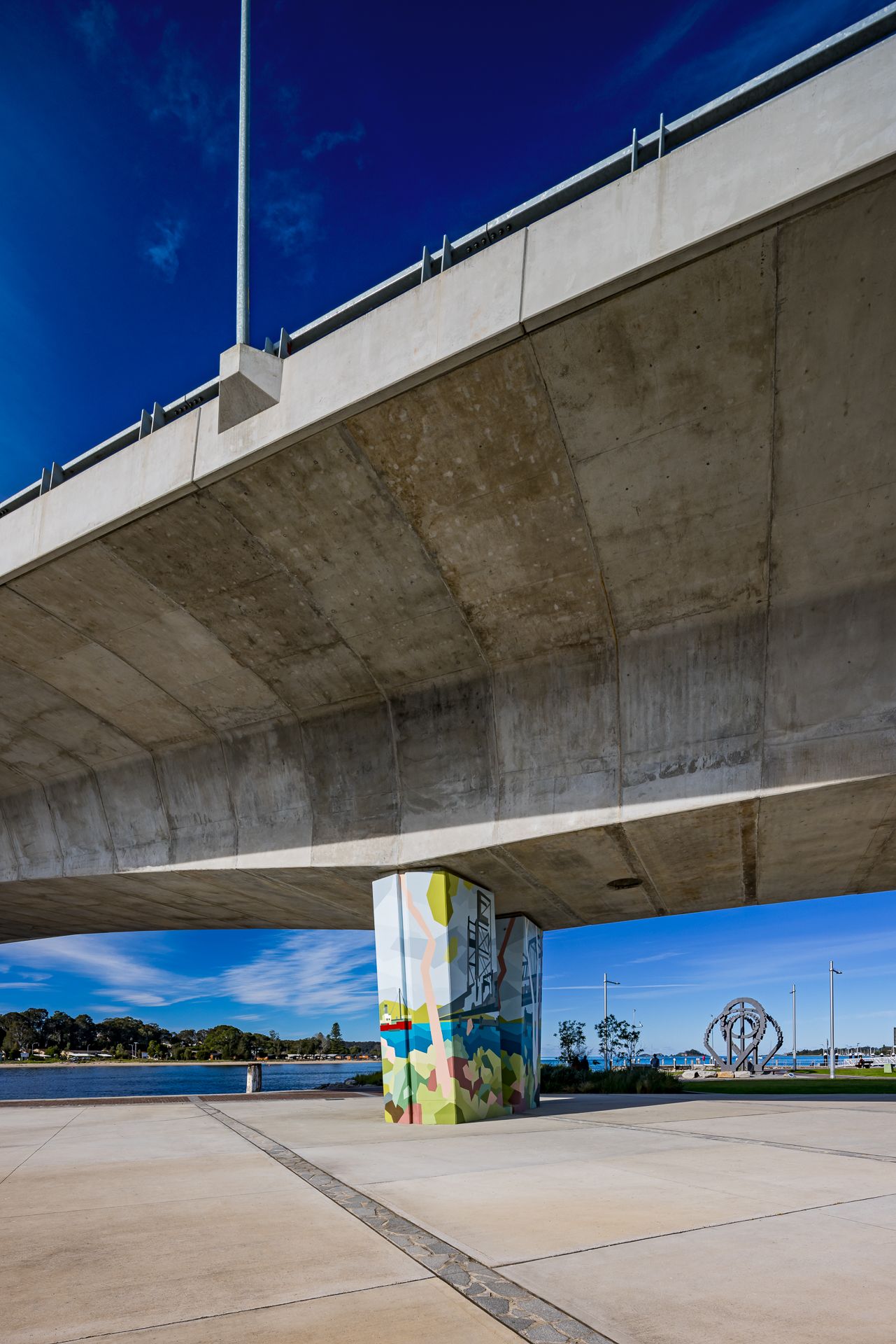 View from beneath a concrete bridge showing a vibrant mural-painted pillar, paved waterfront path and bright blue sky.