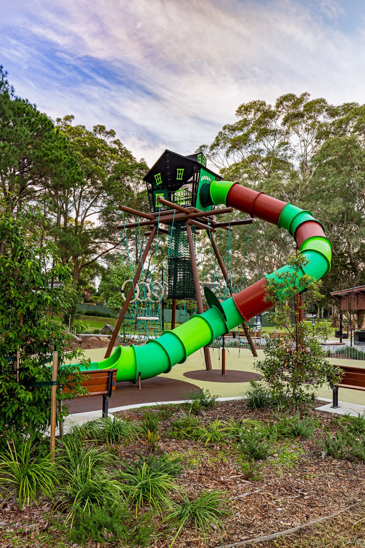 Colorful green and red tube slide on a tall timber climbing tower in a playground surrounded by gum trees and native garden plants.