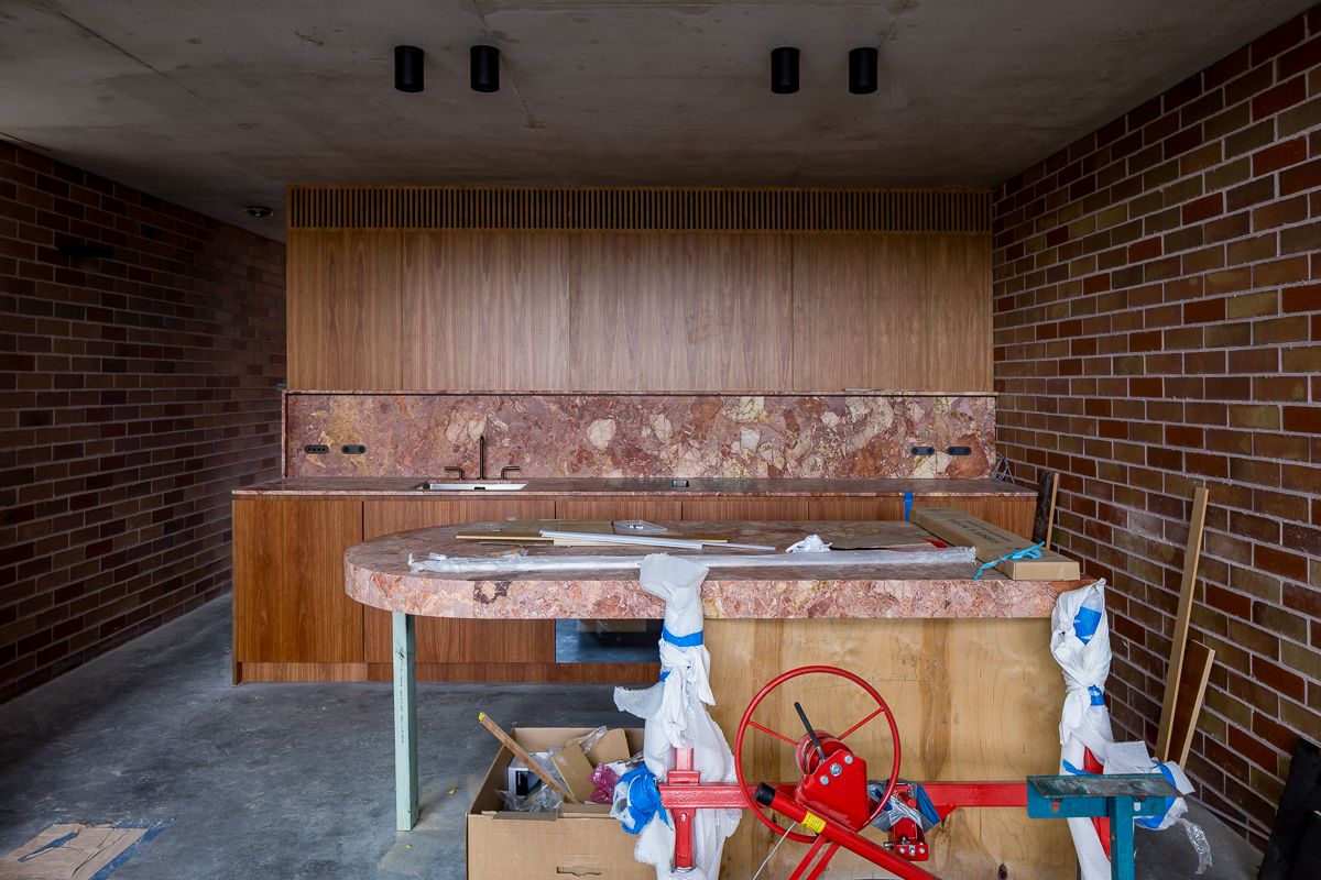 Under-construction kitchen with a red marble splashback, timber joinery, exposed brick walls and a concrete floor.
