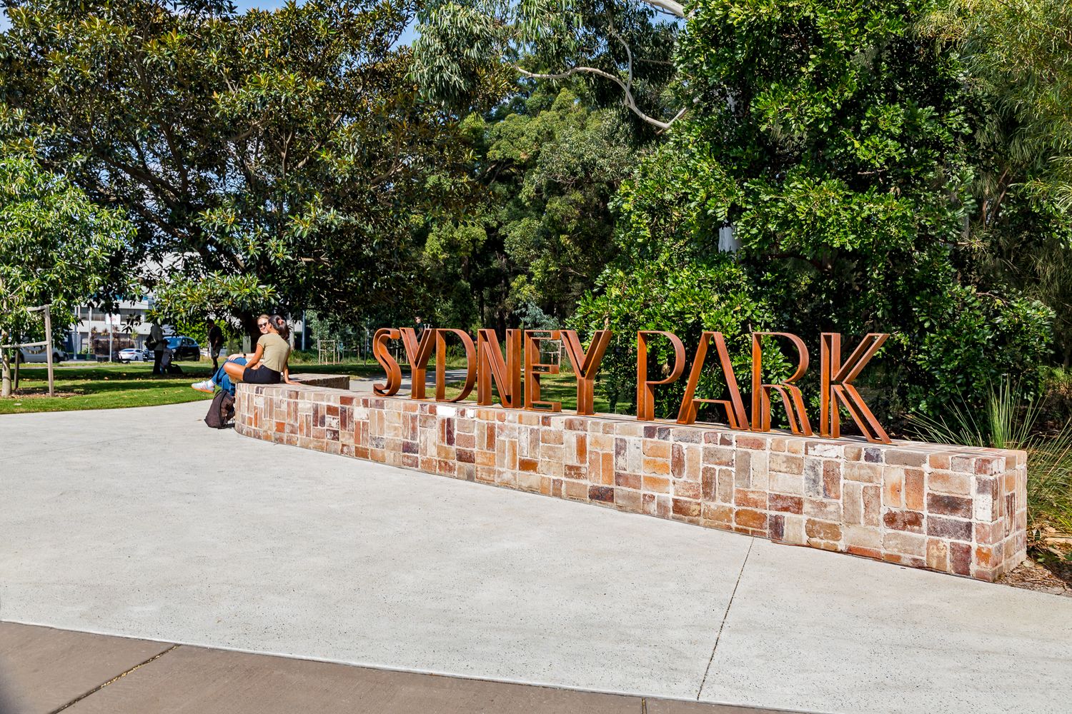 The rusted steel Sydney Park sign stands on a long sandstone wall beside a wide concrete plaza and trees.