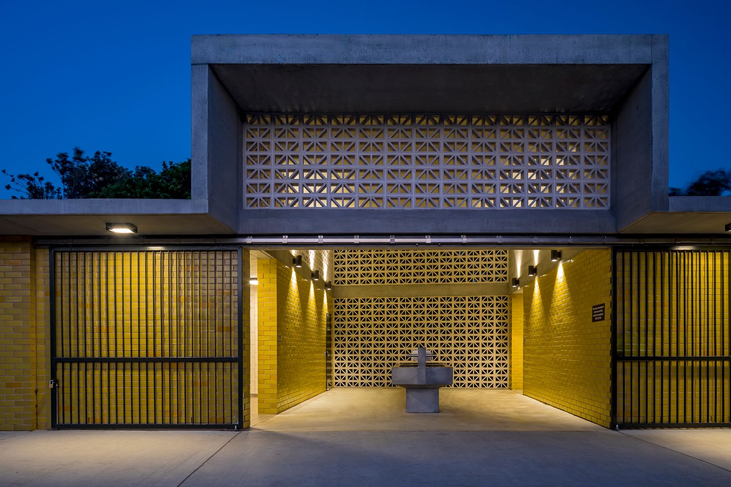 Modern brick building entrance at dusk with a geometric breeze block screen, warm lighting, slatted gates and a stone fountain in the courty