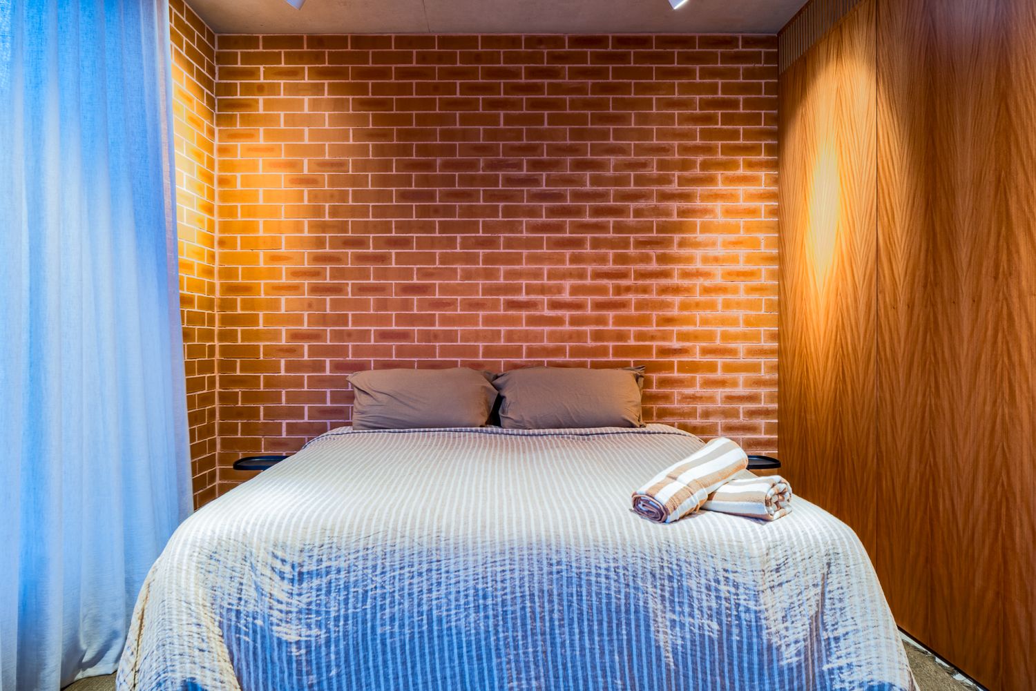 Minimalist bedroom with a bed in blue striped linen against an exposed brick wall, timber panelling, folded towels and blue curtains.