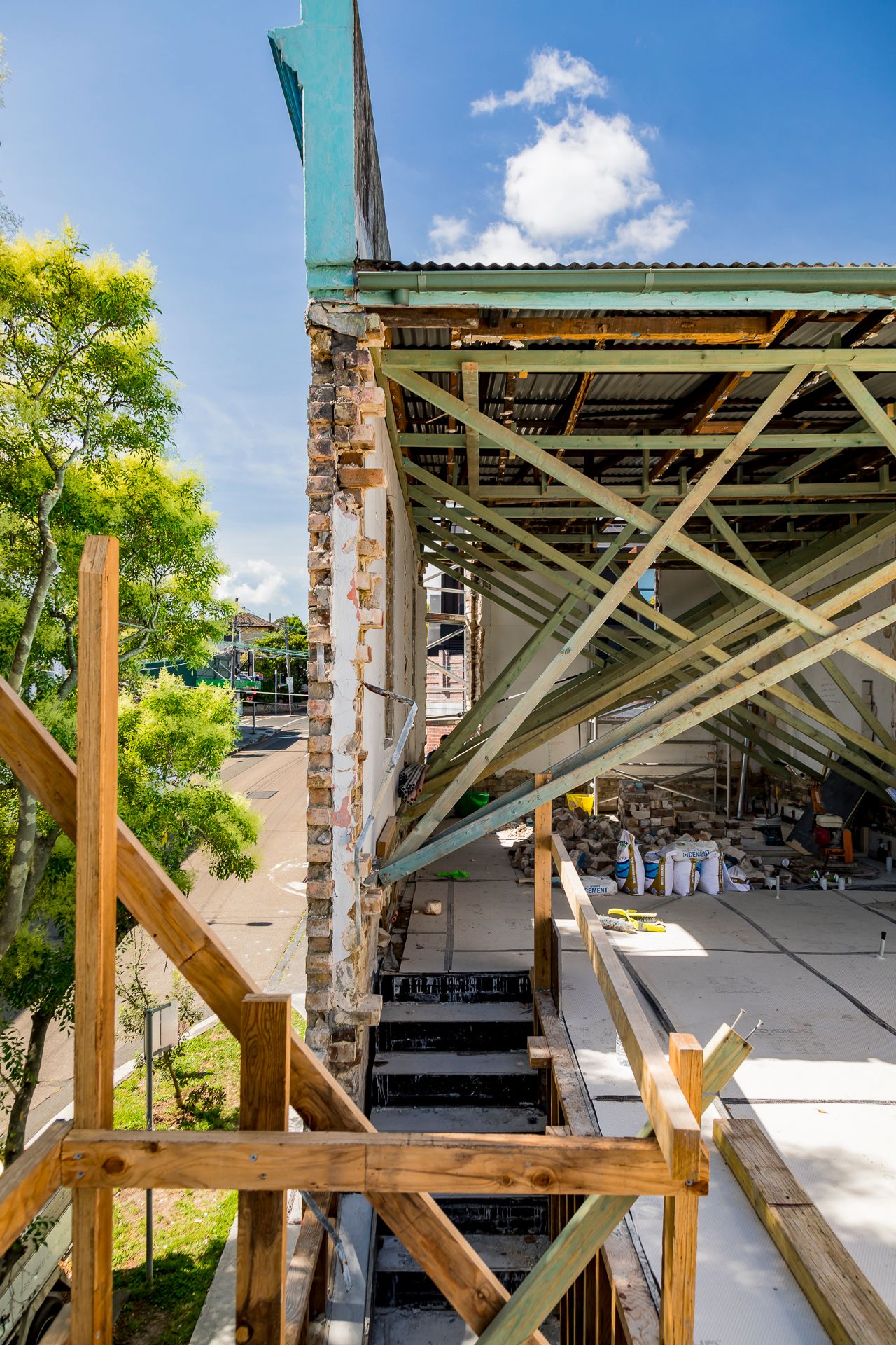 Timber scaffolding and steel roof framing beside an exposed brick wall on a building under renovation against a blue sky.