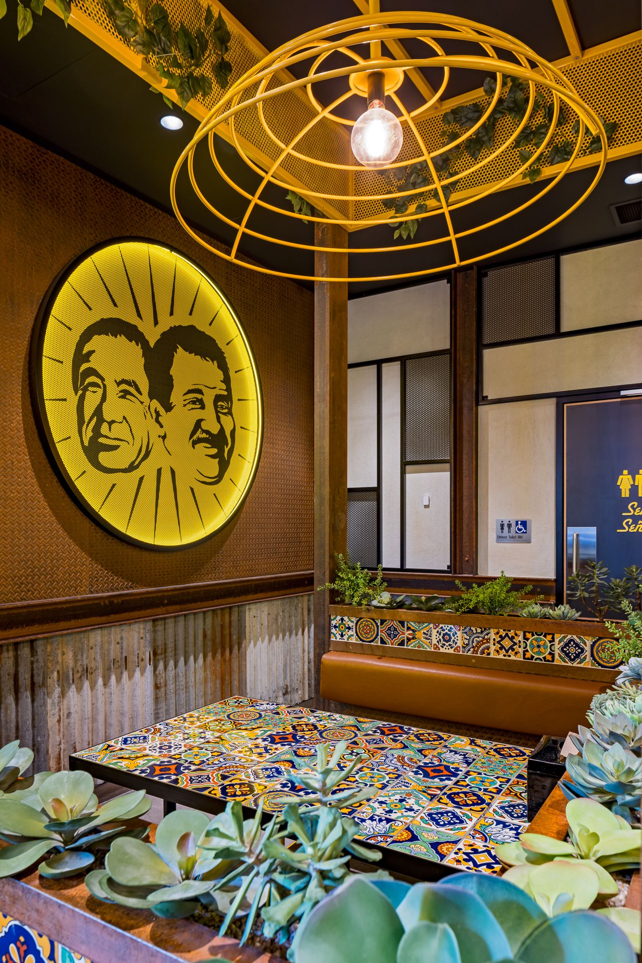Colorful restaurant interior with a round yellow illuminated wall sign, caged pendant light, tiled table, and succulent plants.