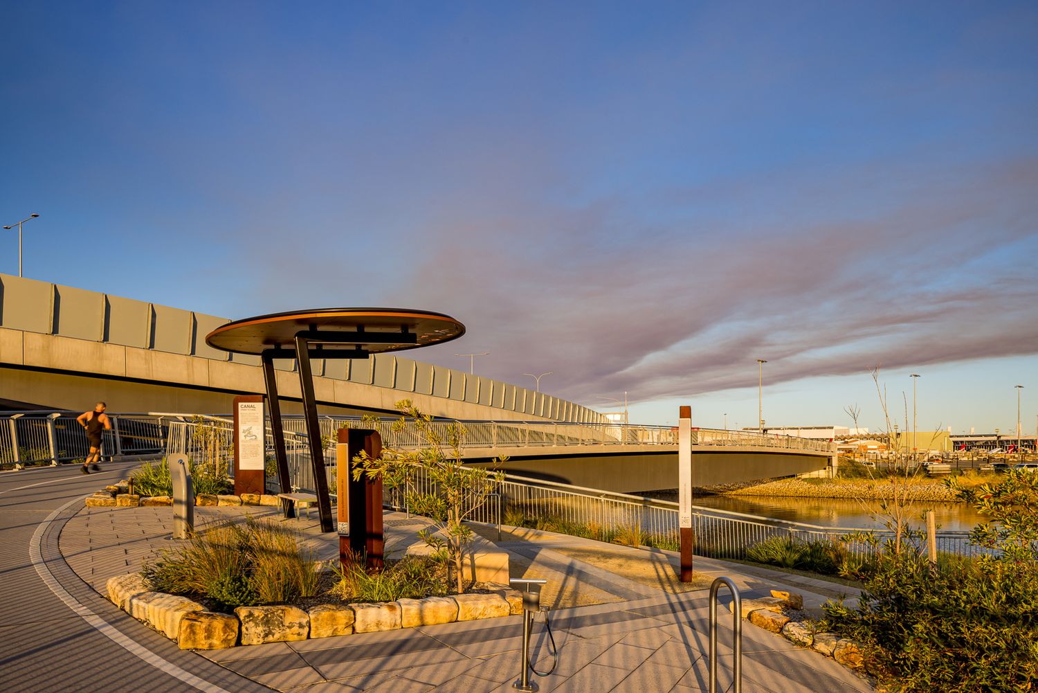 Circular shade canopy and interpretive signs along a curved waterfront path beside an elevated bridge under a dramatic evening sky.