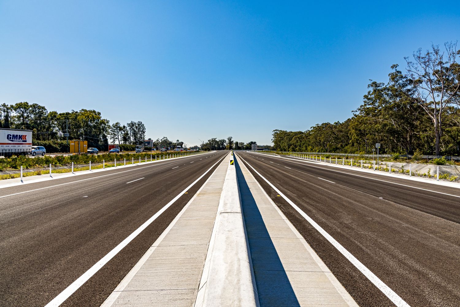 Newly built multi-lane motorway with a concrete median, white line markings and trees lining both sides under clear blue sky.