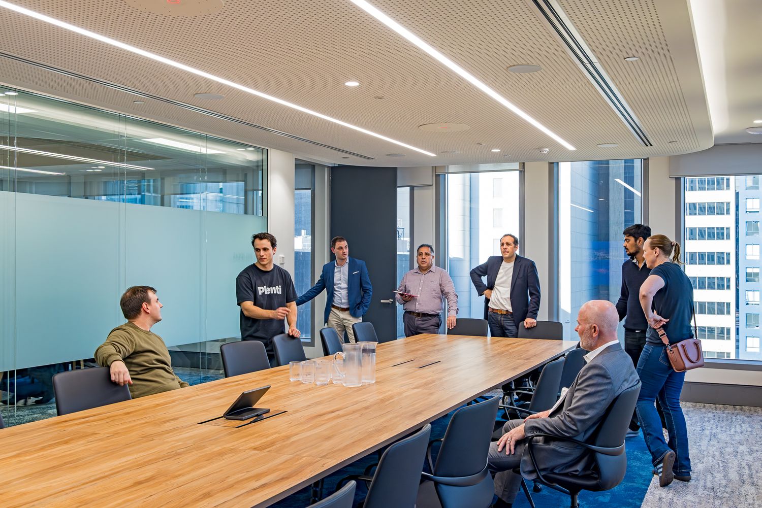 Team members stand and sit around a long timber table in a modern boardroom with floor-to-ceiling windows and city views.