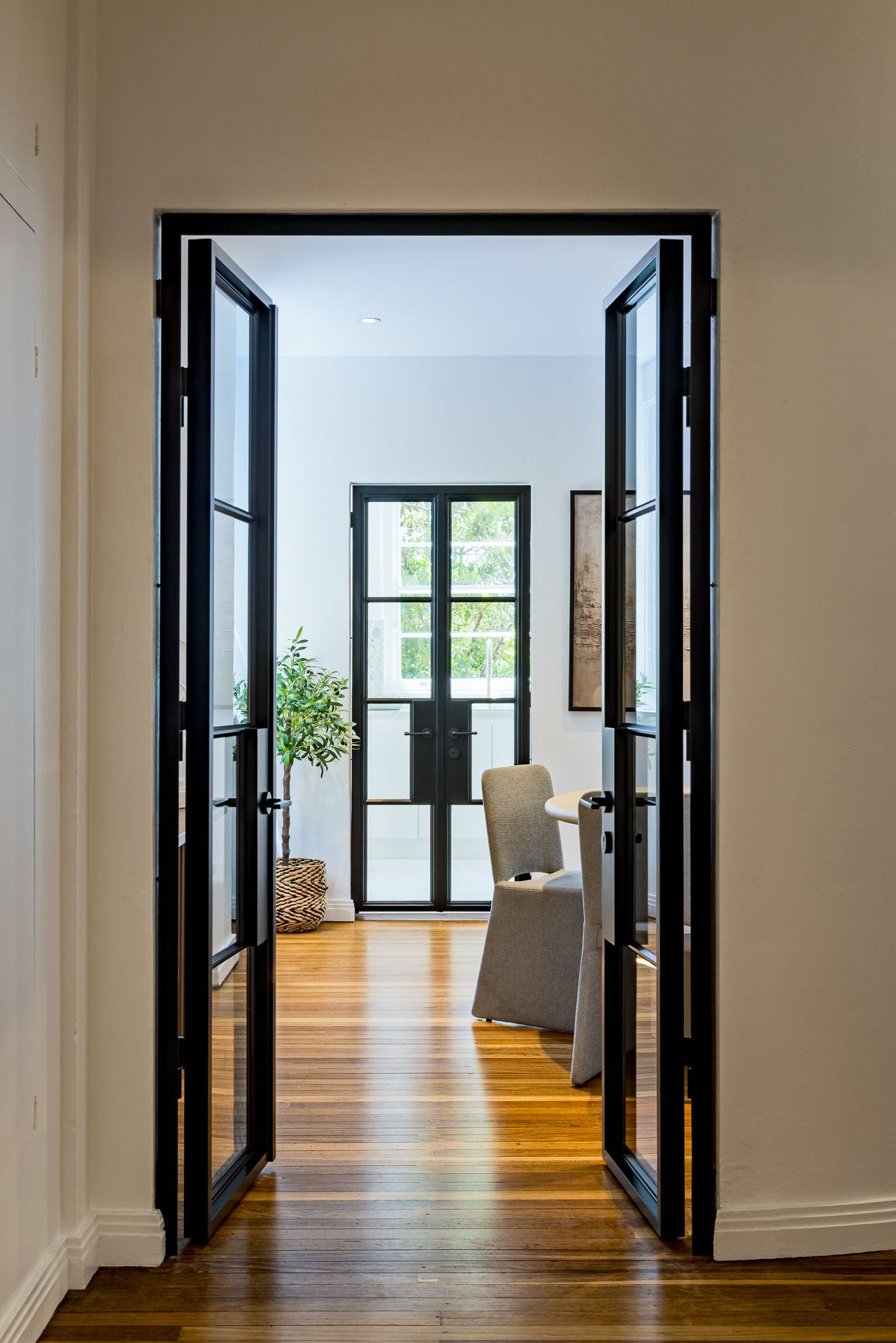 View through open black steel-framed glass doors to a dining room with round table, plant and hardwood floors.
