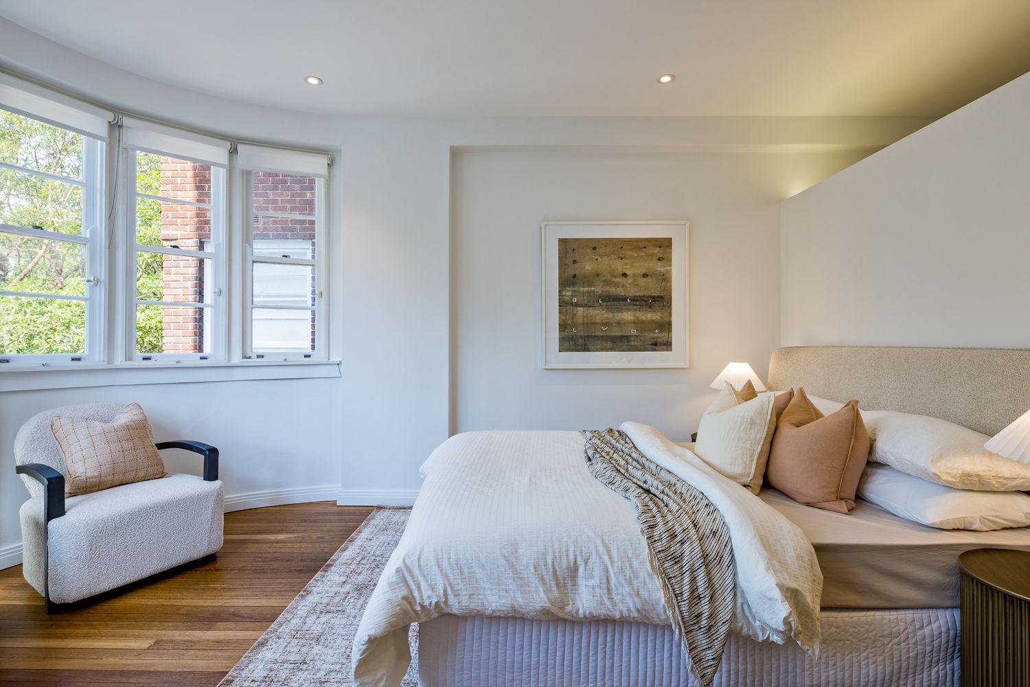Light-filled bedroom with curved bay windows, white bedding, tan cushions, an armchair and an abstract painting above the bed.