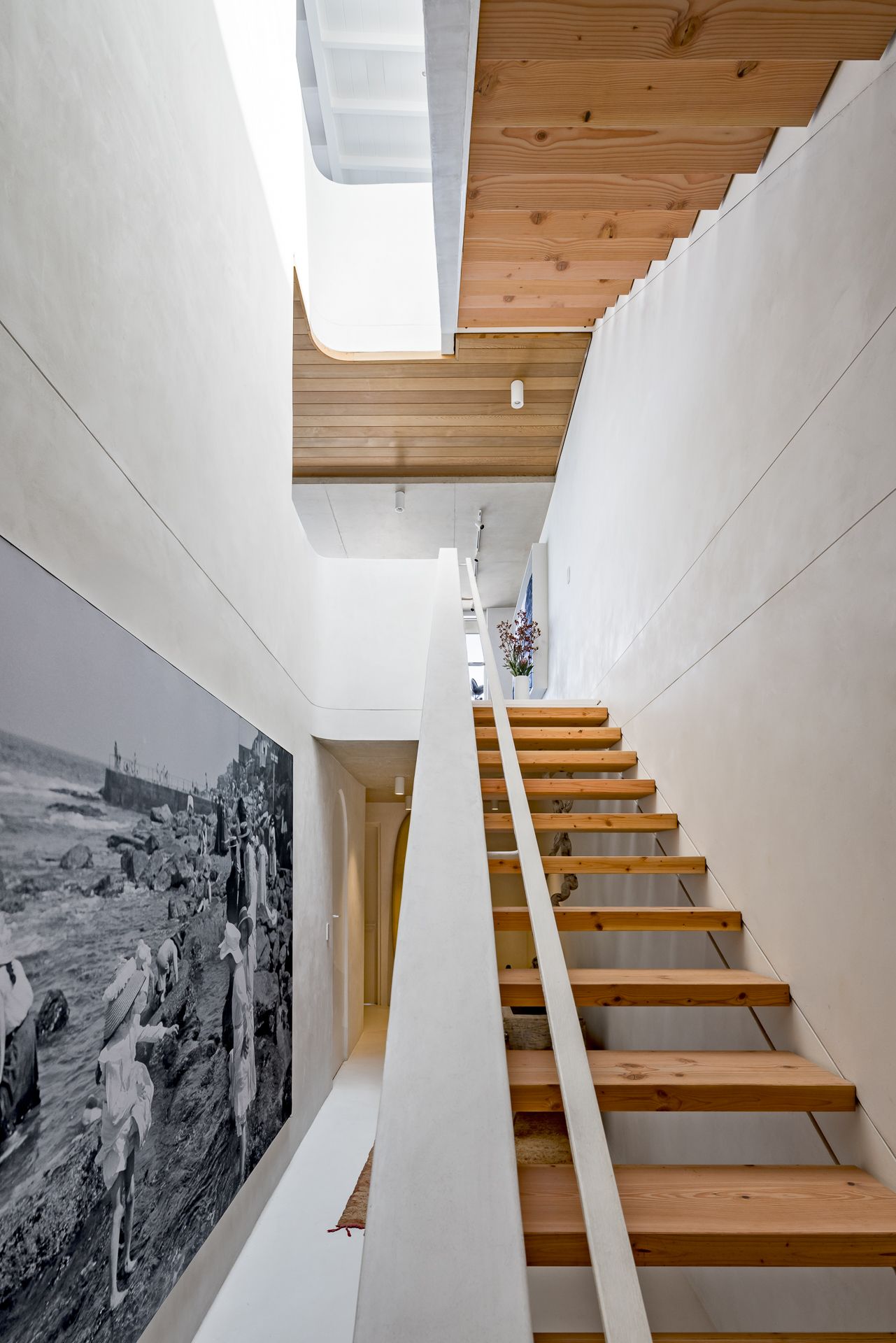 Sculptural timber staircase rising beside a plaster wall with a large black-and-white beach photograph and natural skylight.