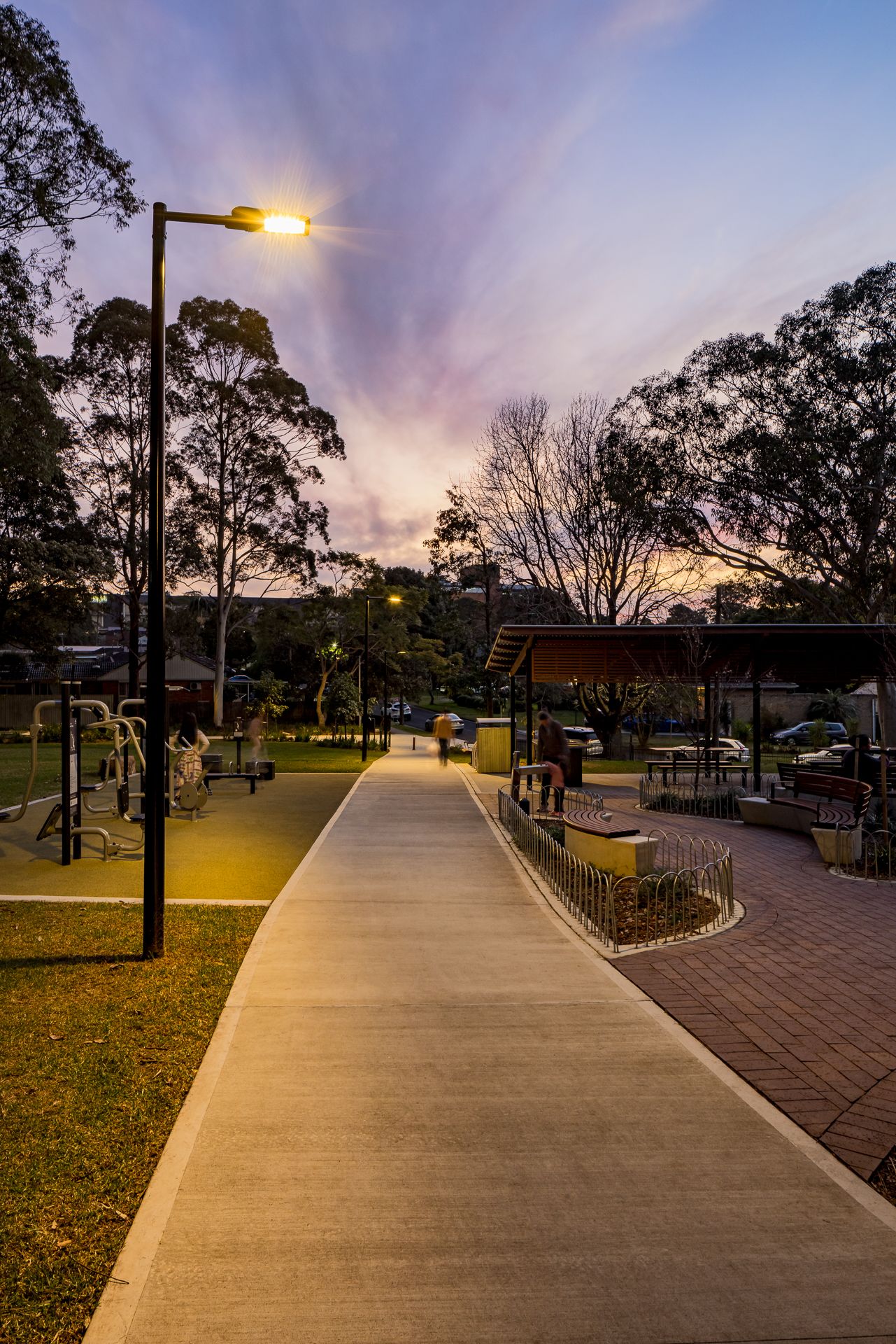 Concrete pathway through a tree-lined park at dusk with a glowing street lamp, outdoor fitness equipment and a covered picnic area.