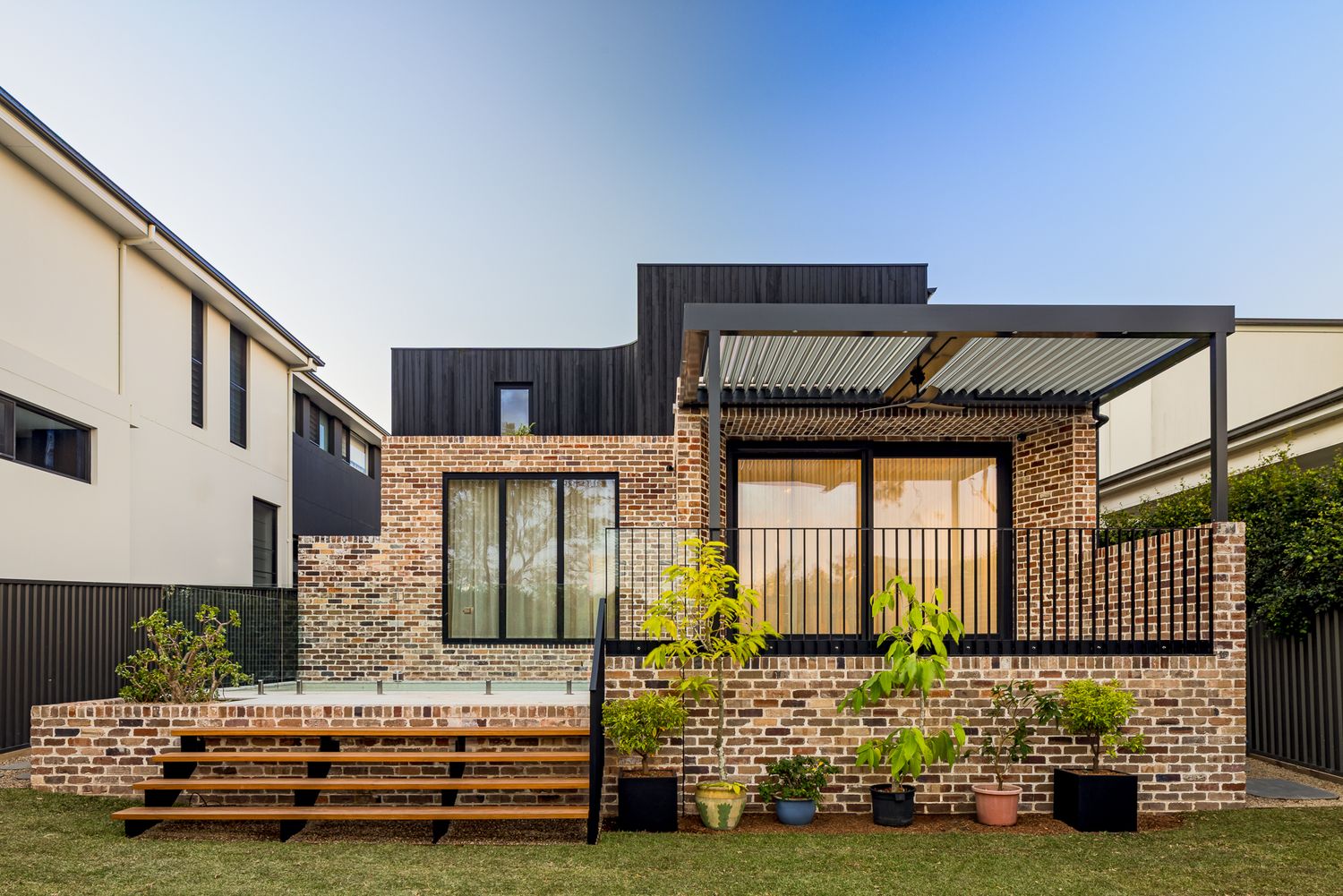 Rear view of a contemporary brick and black timber house with large black-framed windows, pergola, raised deck and potted plants.