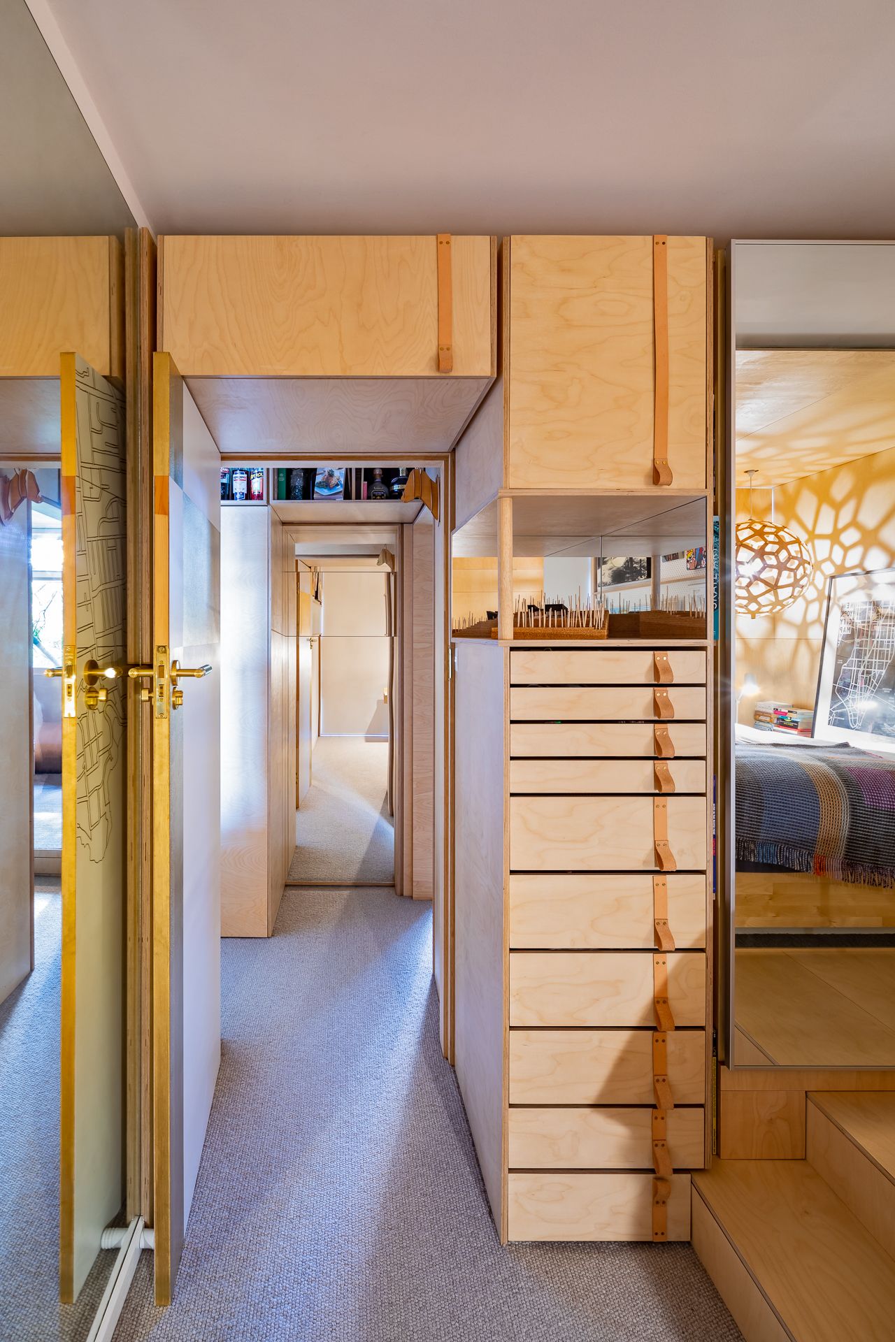 Narrow hallway with plywood cabinets, leather-handled drawers, brass door handles, and a view into a bedroom.