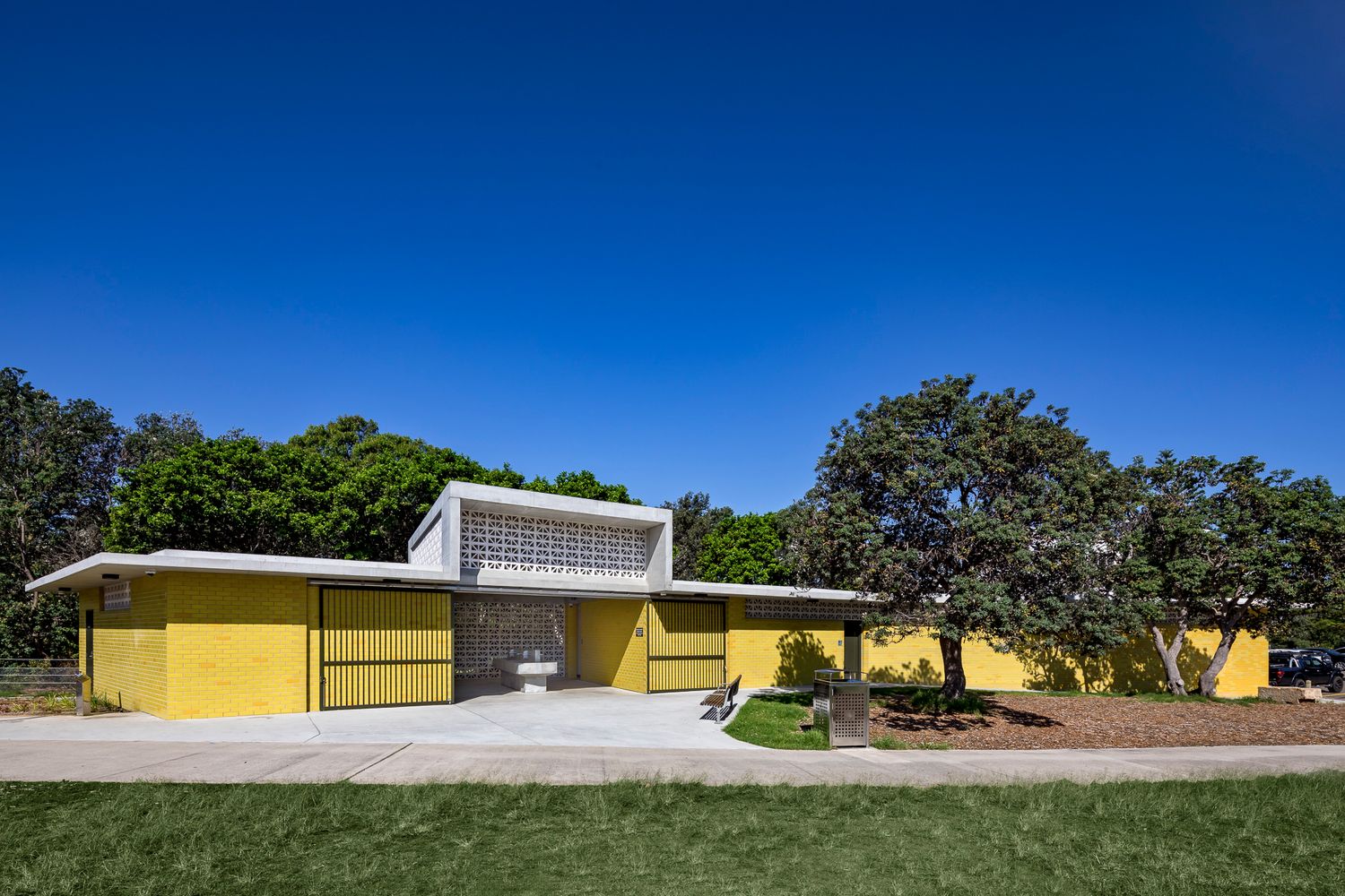Front view of a modernist yellow brick park building with a raised concrete clerestory and breeze block detail beside leafy trees.